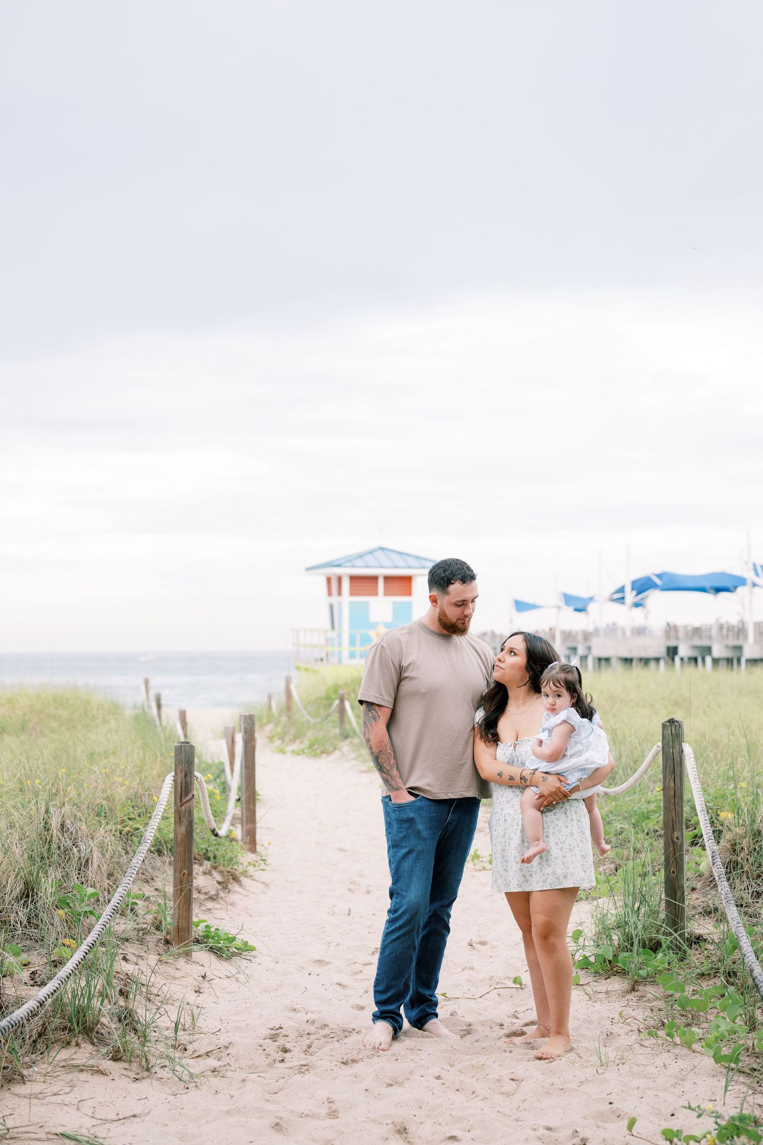 A couple walks hand in hand along a sandy beach path bordered by grass and wooden posts on a sunny day.