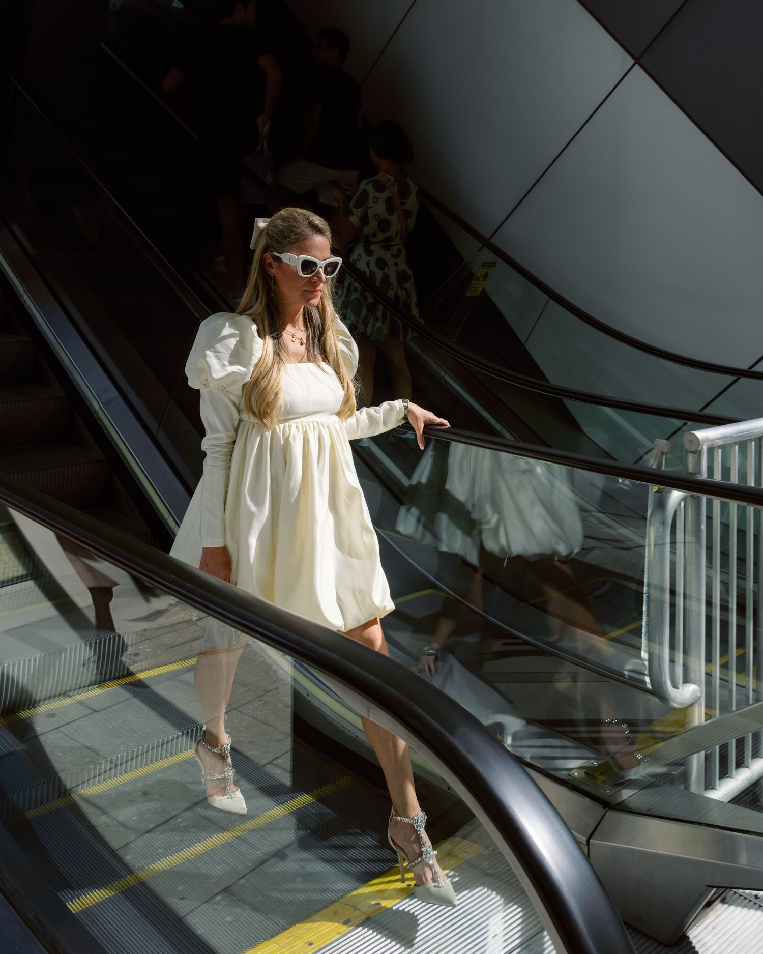 A sequence showing someone in a white dress descending an escalator in a modern shopping mall or building.