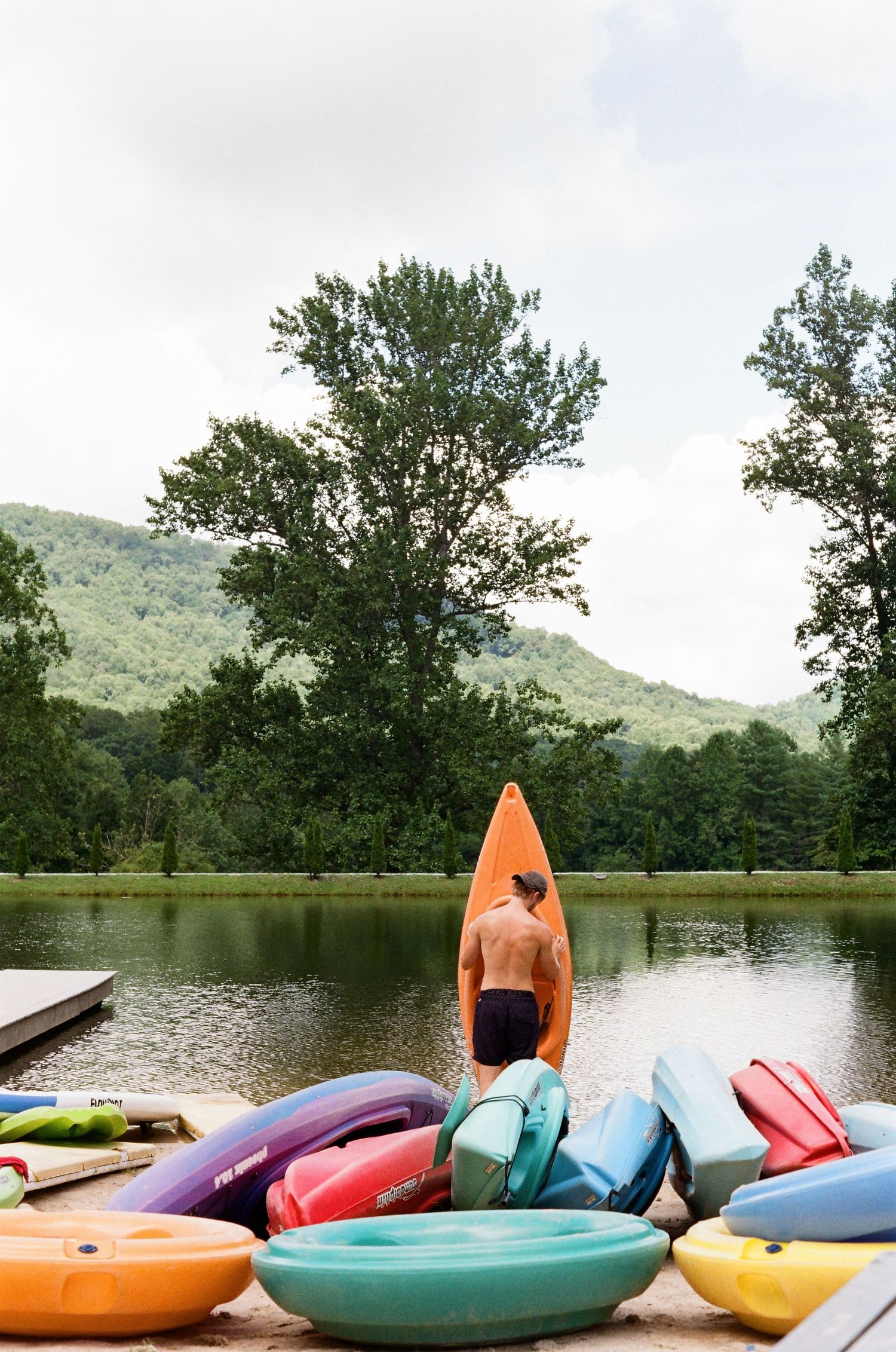 Shirtless man carrying an orange kayak toward a calm lake surrounded by colorful kayaks and lush green mountains.