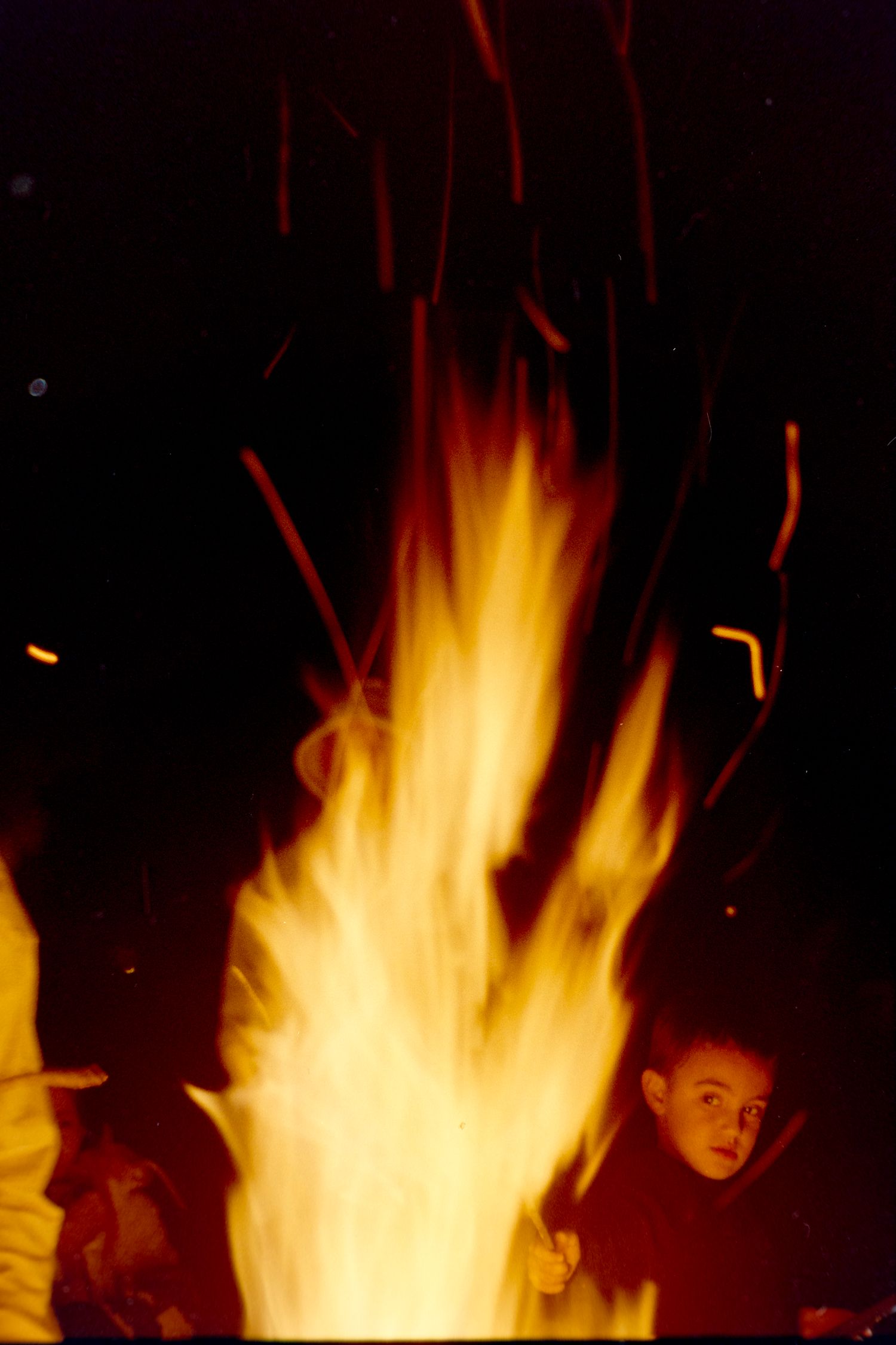 A child's face glows in the light of a large campfire with bright yellow flames and sparks flying into the dark night sky.