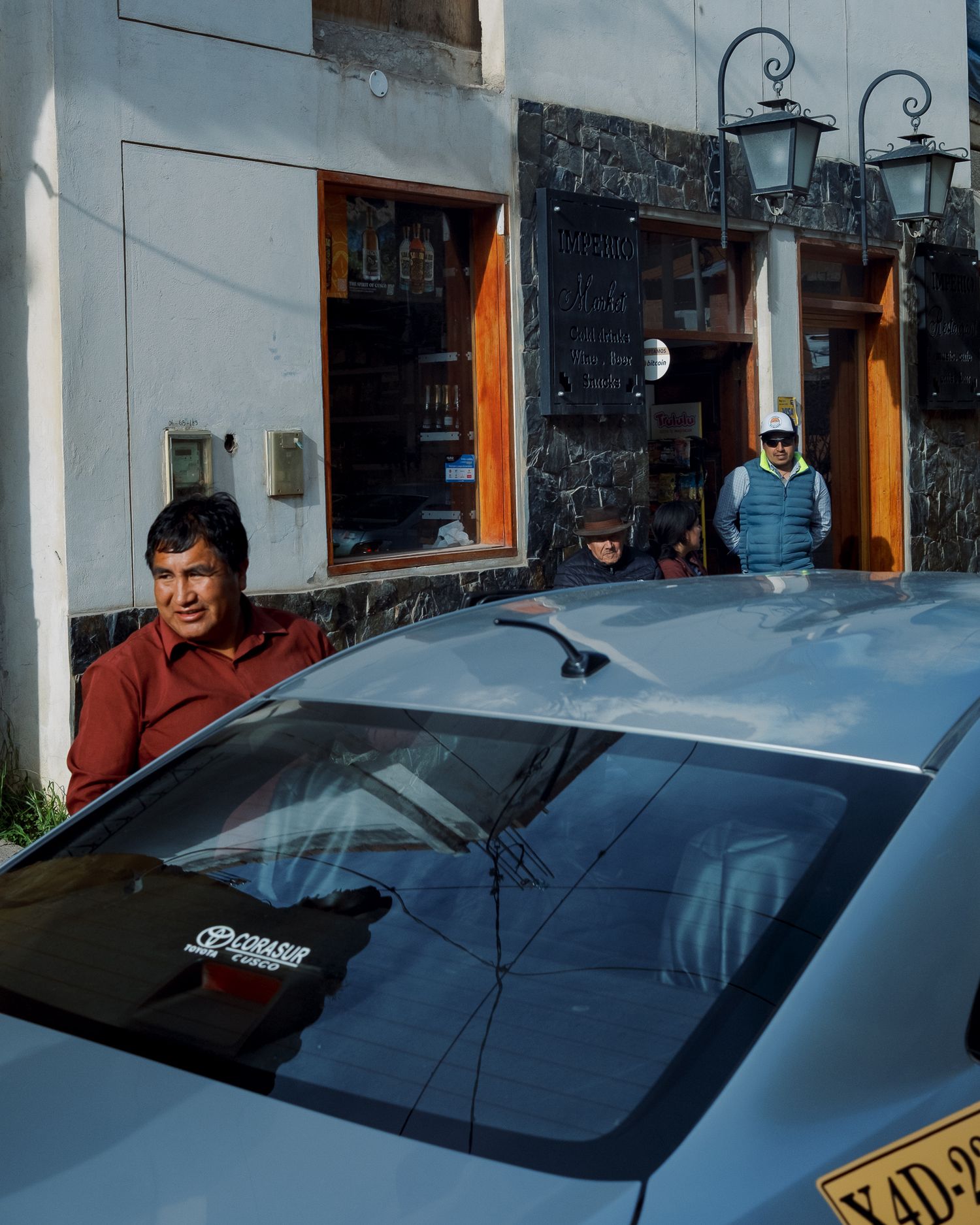 Three men stand near a blue car outside a rustic shop with orange doors and stone walls in a Latin American town.