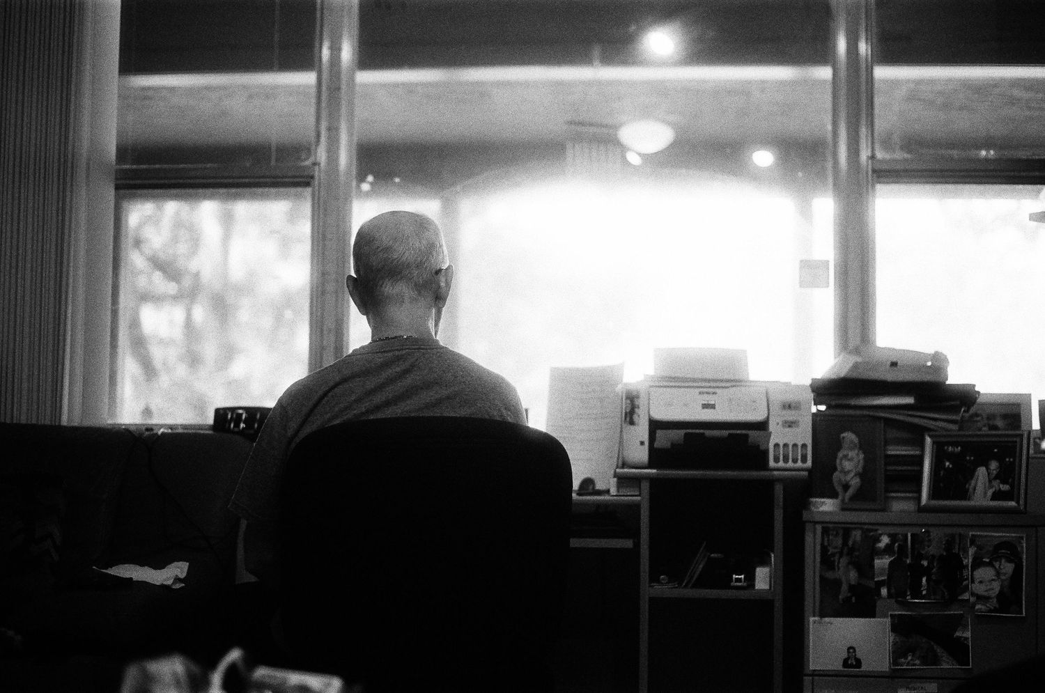Black and white photo of a bald person sitting at a desk with a printer, facing large windows in a dimly lit room.