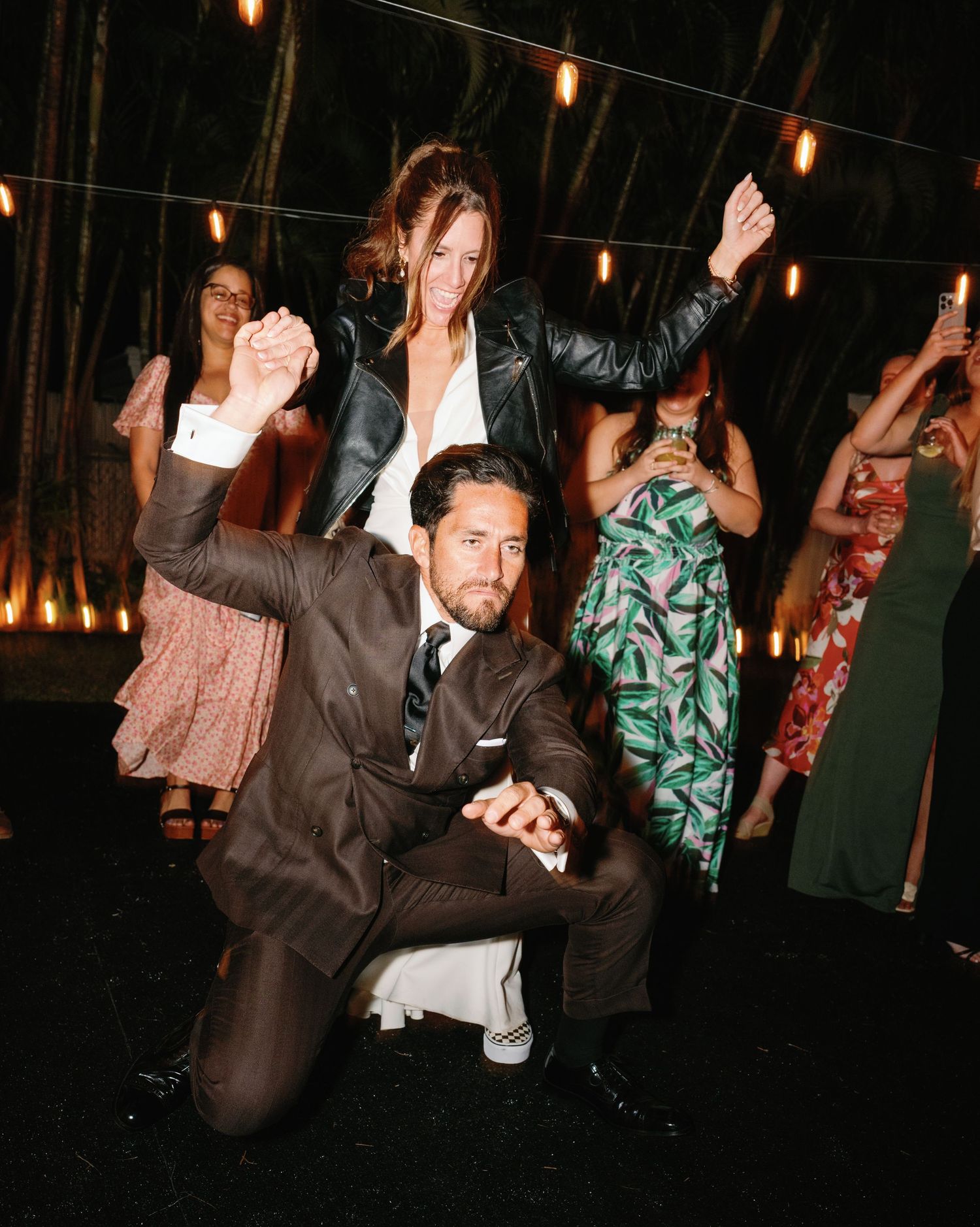 Wedding guests dance and celebrate on a dimly lit dance floor with string lights overhead.