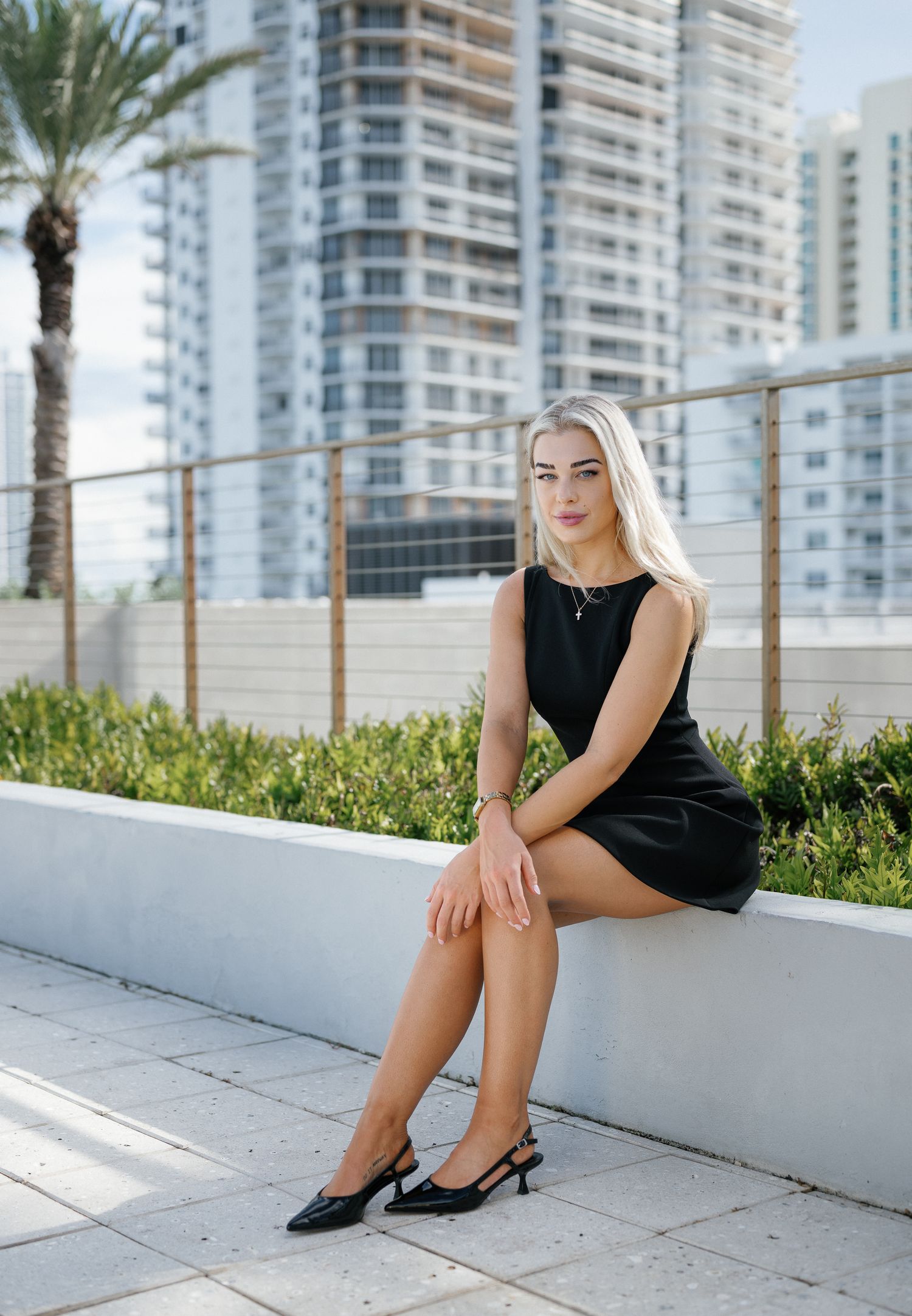 A model in a black dress sits on a white concrete ledge with modern high-rise buildings and palm trees in the background.