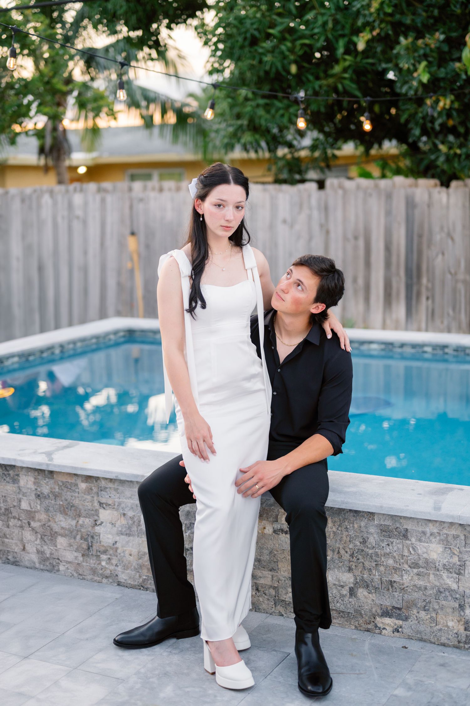 A couple poses together by a swimming pool with a wooden fence backdrop.