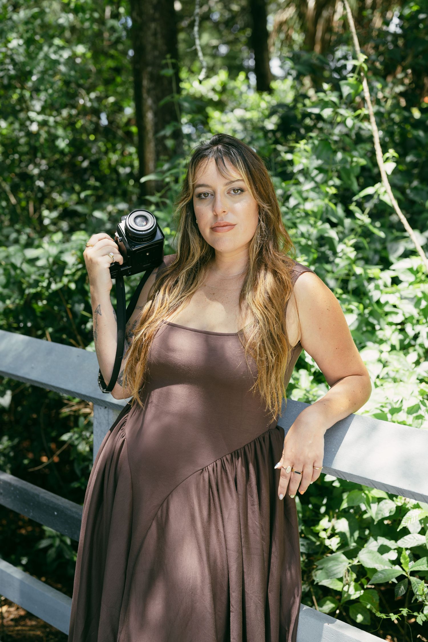 A model in a brown dress poses outdoors against lush green foliage in a natural light photoshoot series.