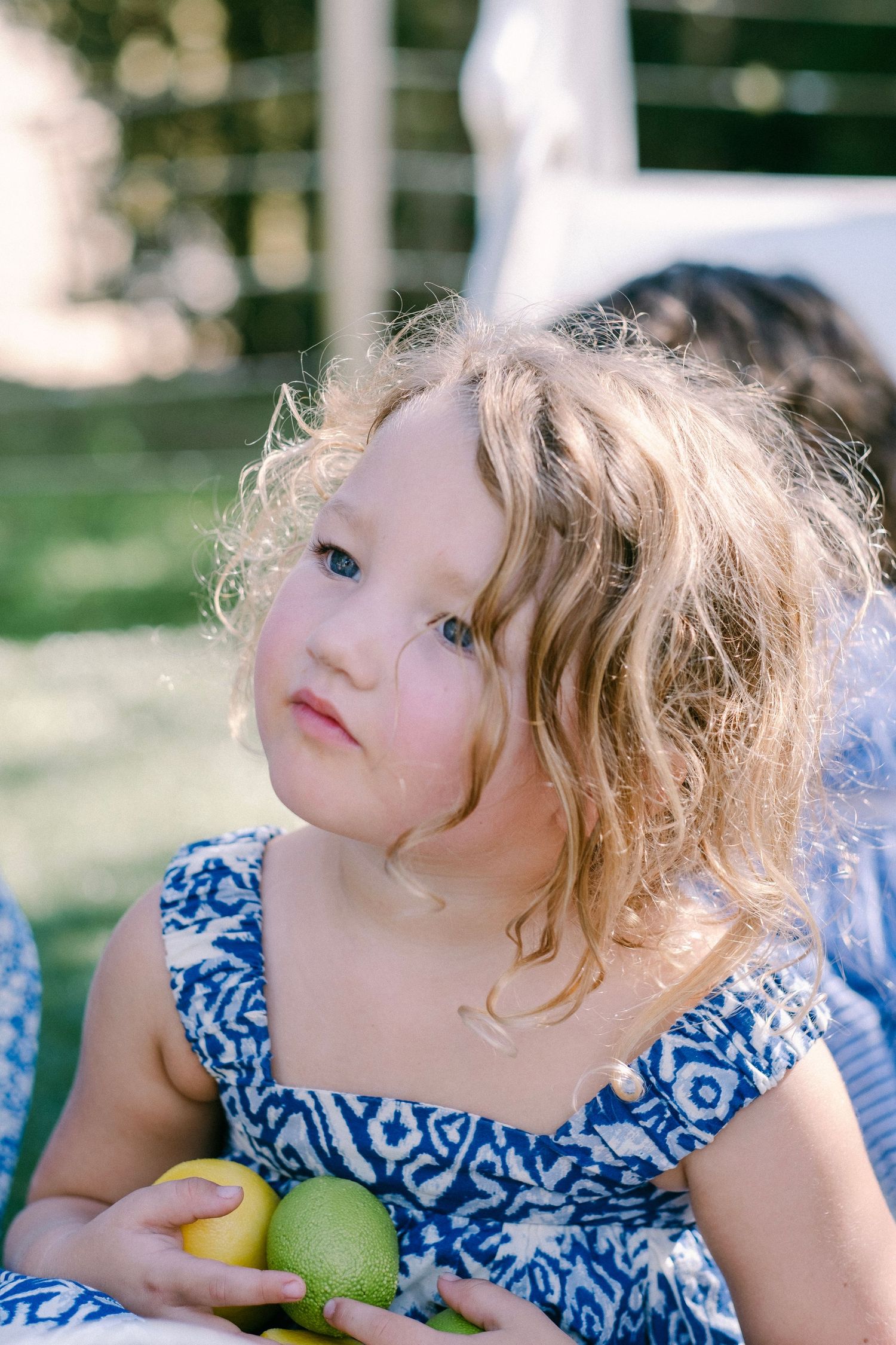 Young child in a blue and white patterned dress holds a green apple while looking upward on a sunny day.