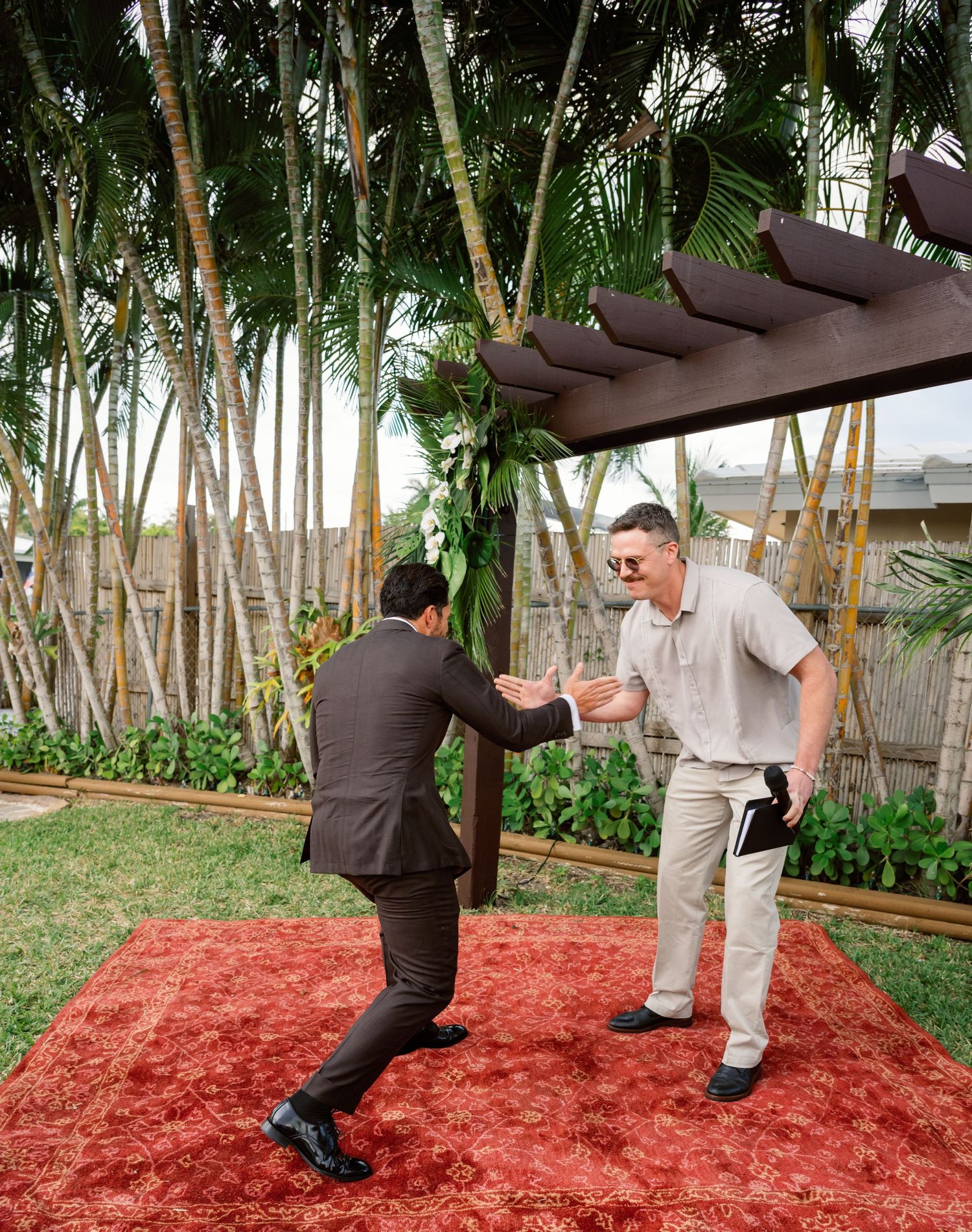 Two people practicing martial arts moves on red mulch ground surrounded by tropical palm trees.