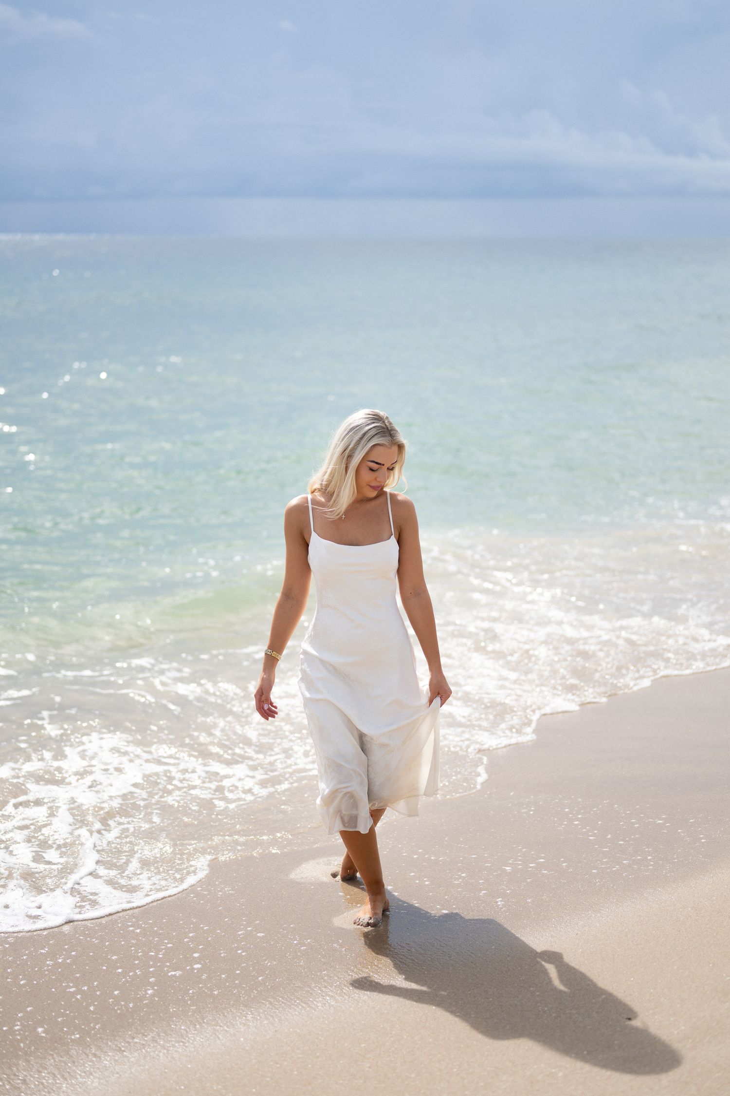 Person in white dress walking along a sandy beach shoreline with gentle waves and soft sunlight.