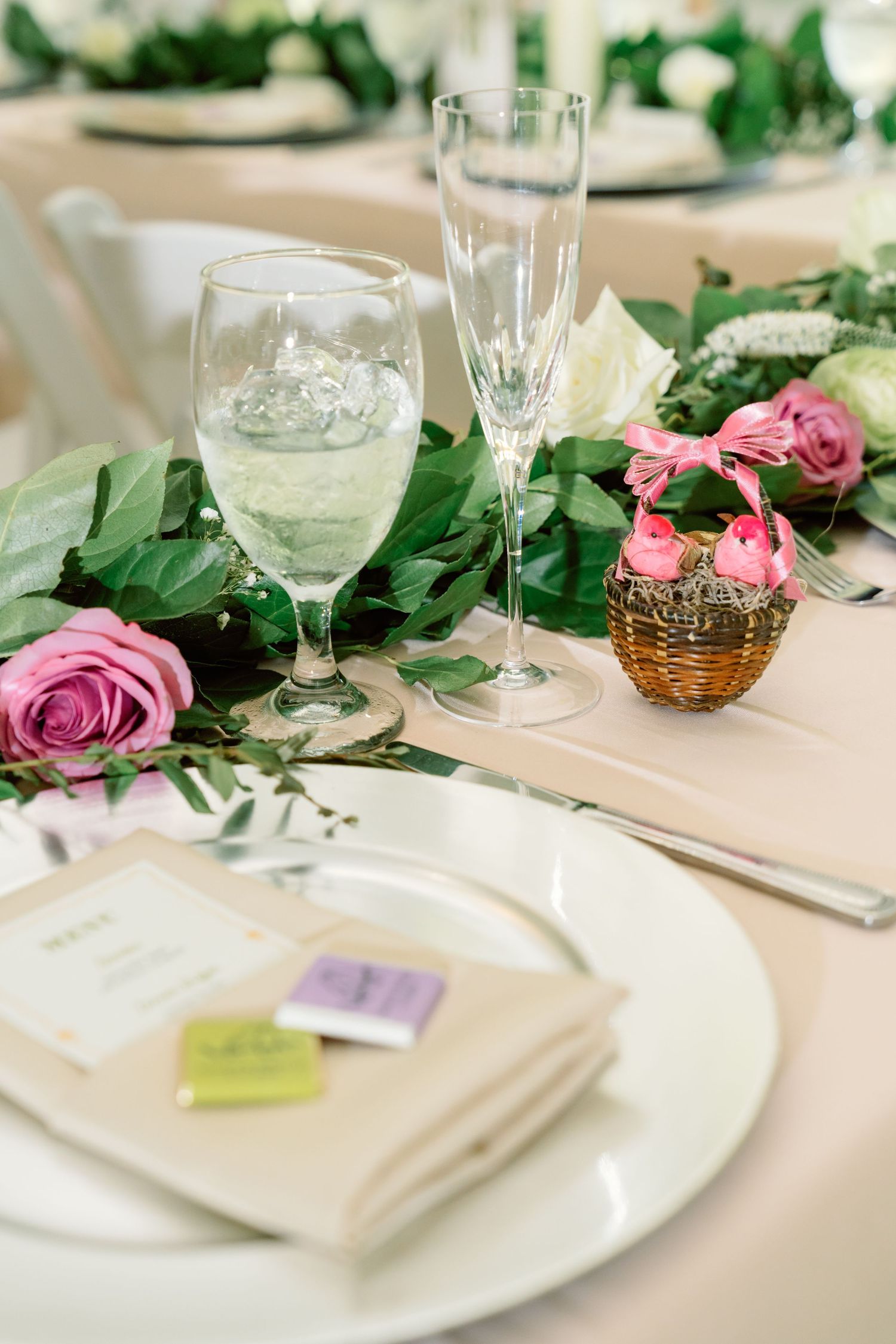 Elegant table setting with wine glasses, pink roses, and green foliage on a white tablecloth.