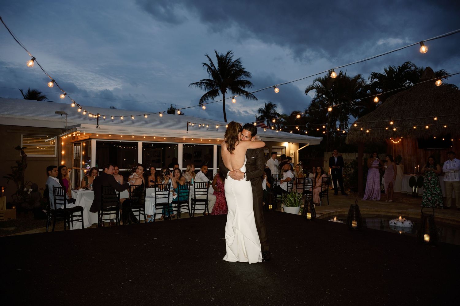 Evening wedding reception under string lights with palm trees silhouetted against a dusky blue sky.