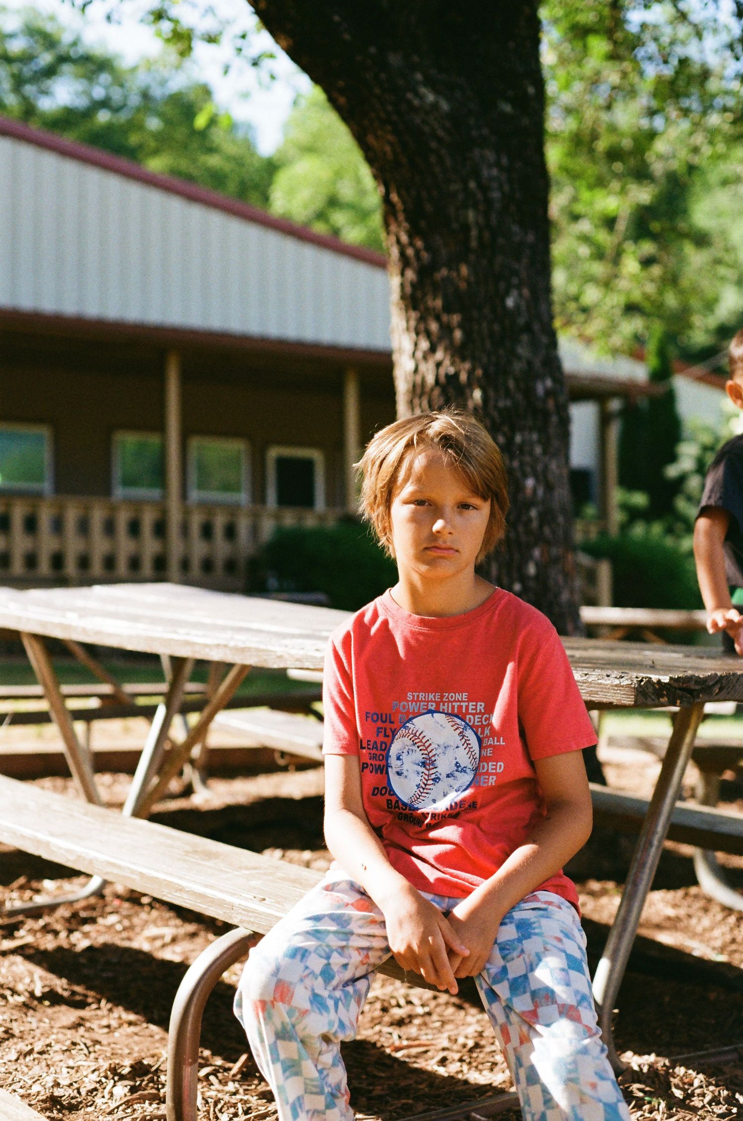 A child in a red t-shirt and patterned pants sits on a wooden picnic table outside a building on a sunny day.