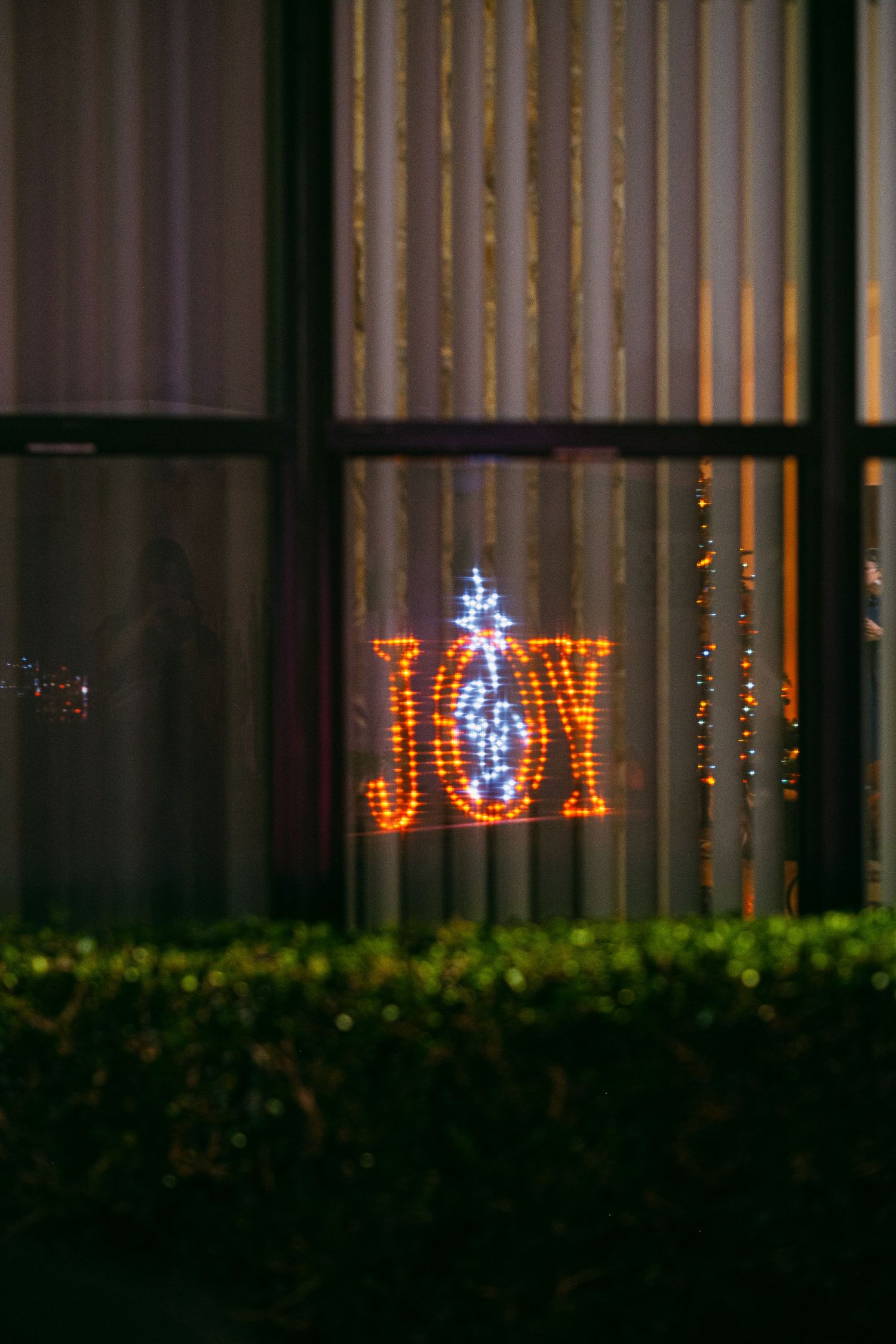 Illuminated holiday decorations spelling 'JOY' displayed through window blinds at night.