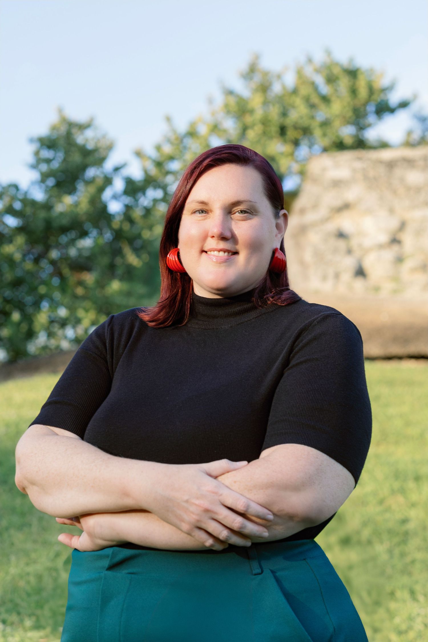 Person wearing black top and teal pants poses outdoors against natural landscape with trees and hills in background.