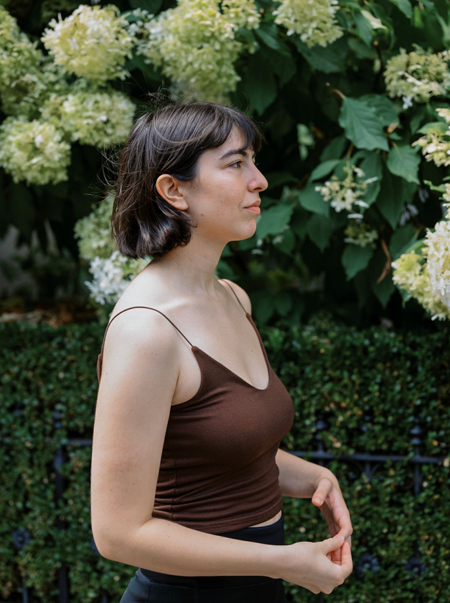 Person in brown sleeveless top poses expressively against backdrop of cream-colored hydrangea blooms and green foliage.