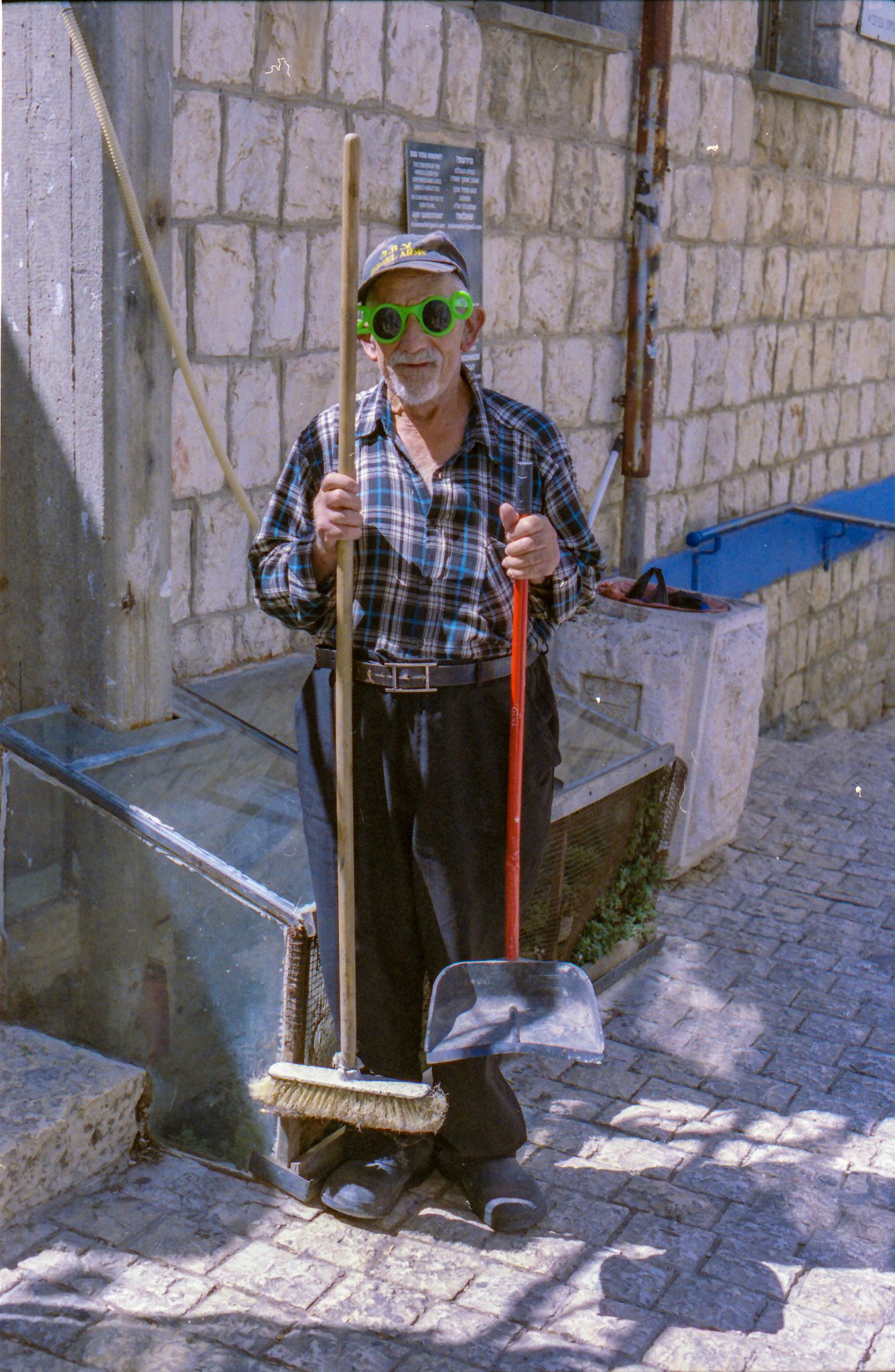 Elderly man holding a broom and dustpan, wearing green novelty glasses and plaid shirt, standing outdoors near a stone building.