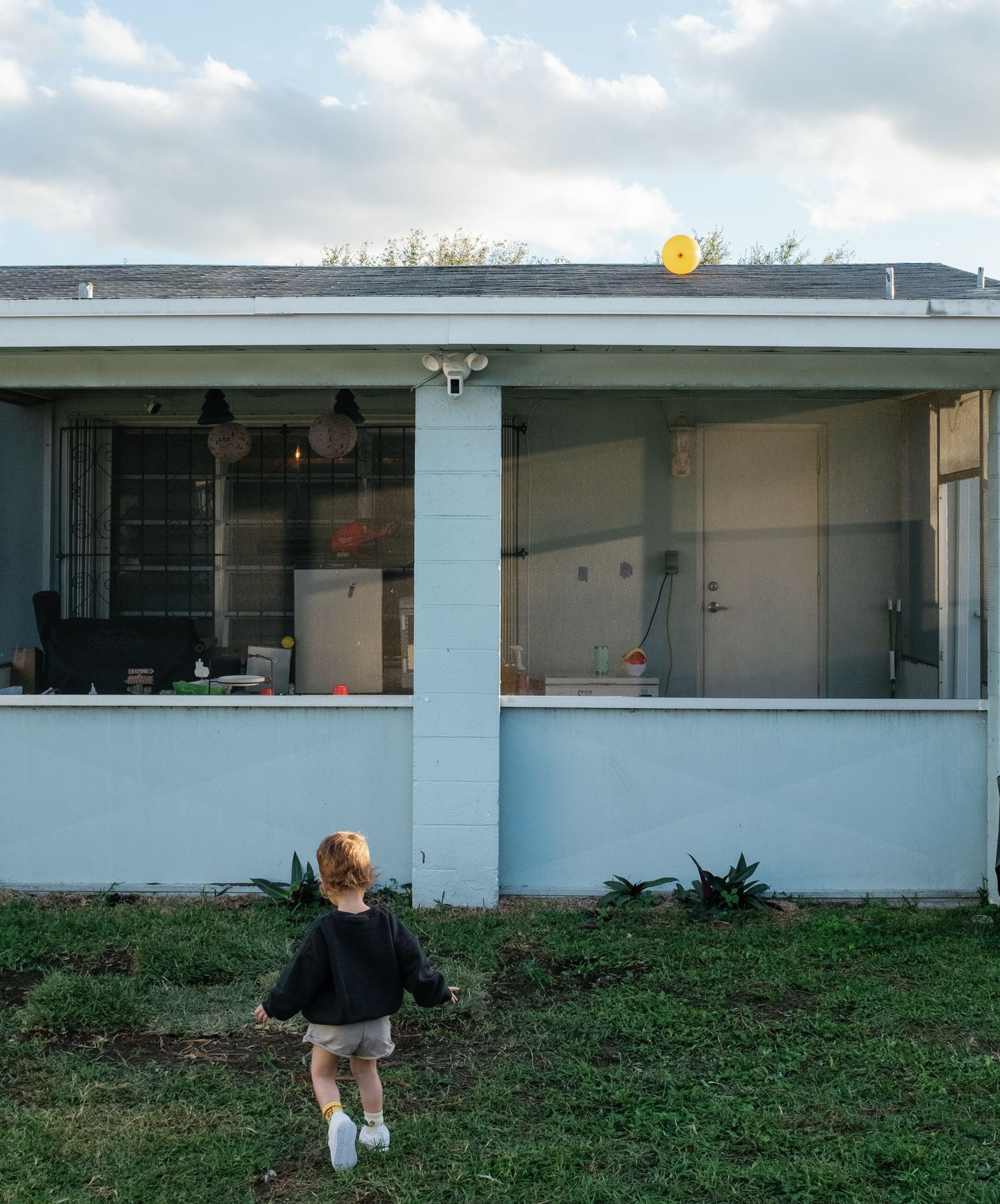 A young toddler with red hair walks toward a light blue house with a screened porch on a grassy lawn.