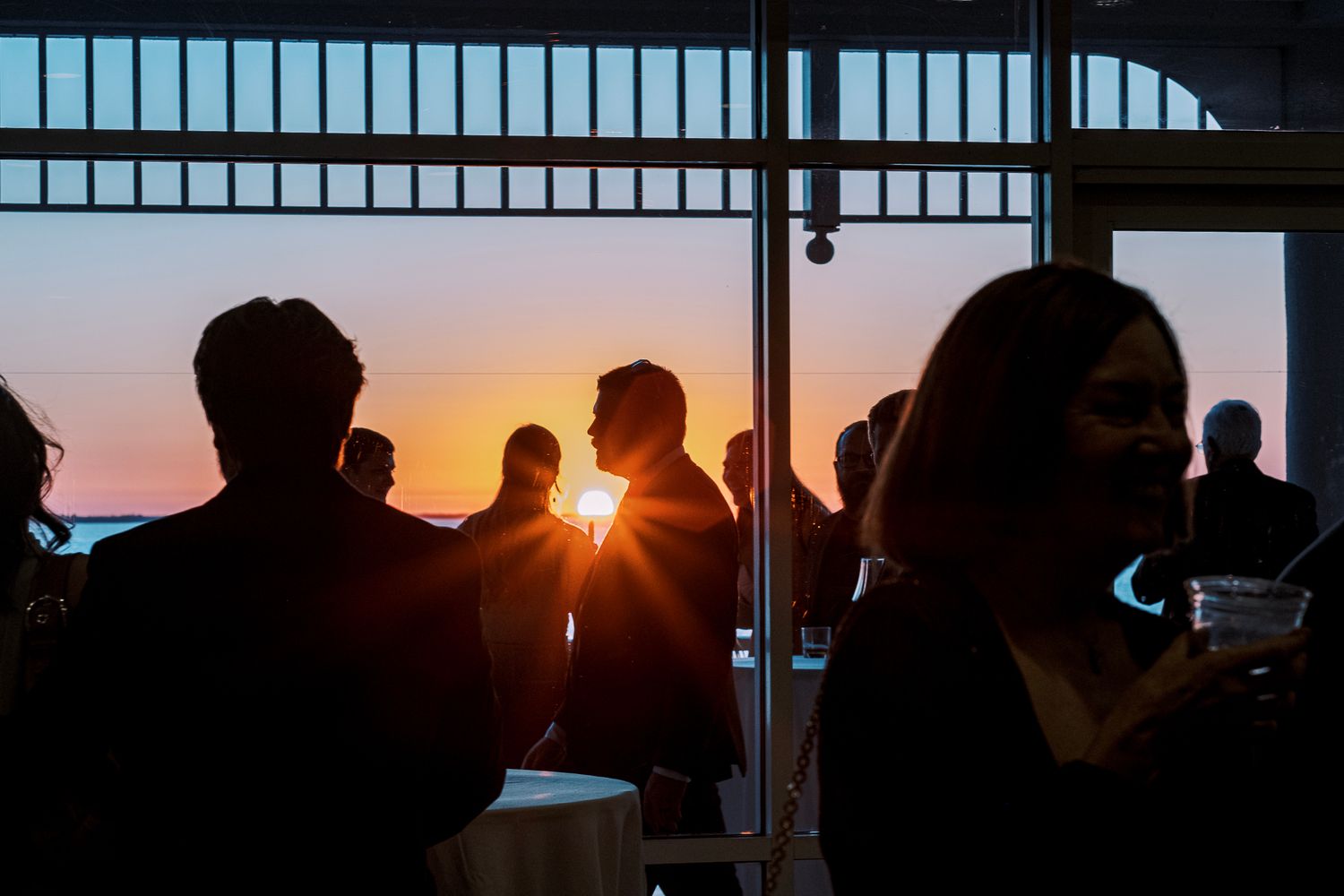 Silhouettes of people gathered at a sunset viewing area with sun rays peeking through vertical window frames.