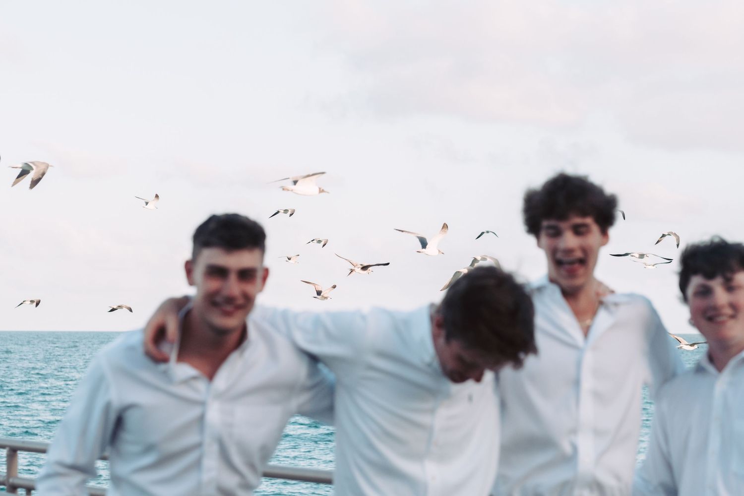 A group of people in white shirts stand together laughing and smiling on a beach with ocean waves in the background.