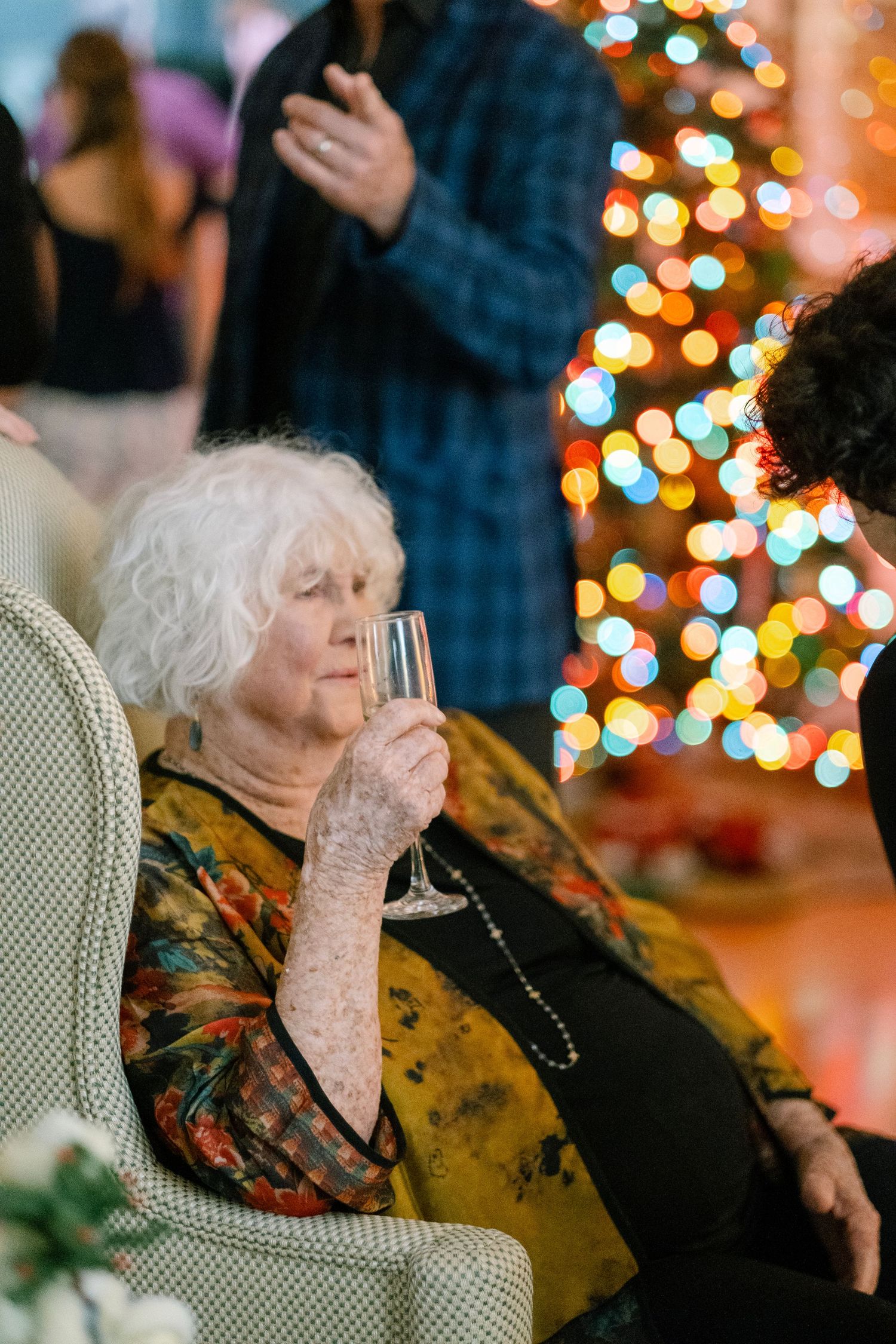 A person in a floral dress sits in a cream colored chair with Christmas lights twinkling in the background.