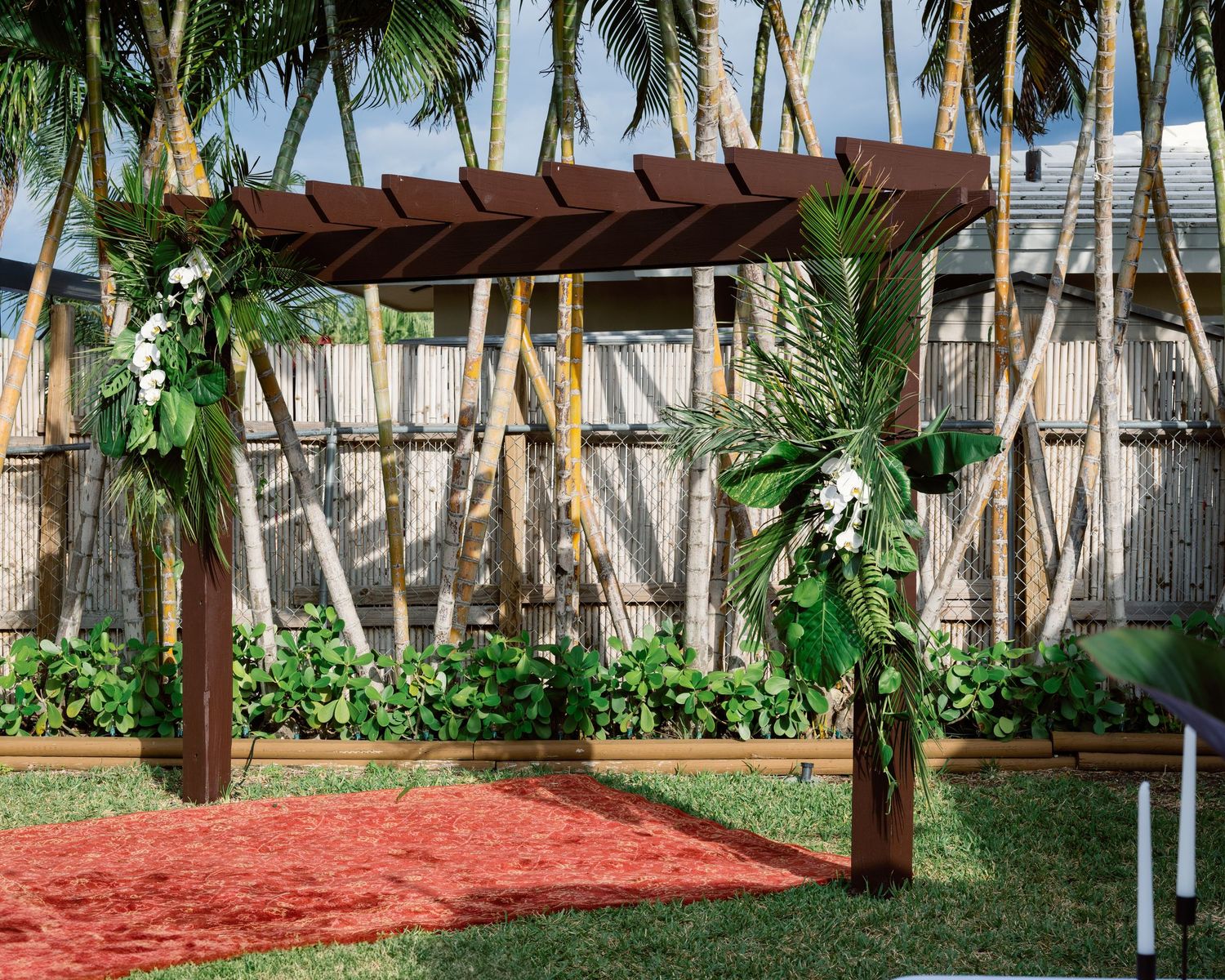 Outdoor wedding venue decorated with palm trees and a wooden pergola on a grassy lawn.