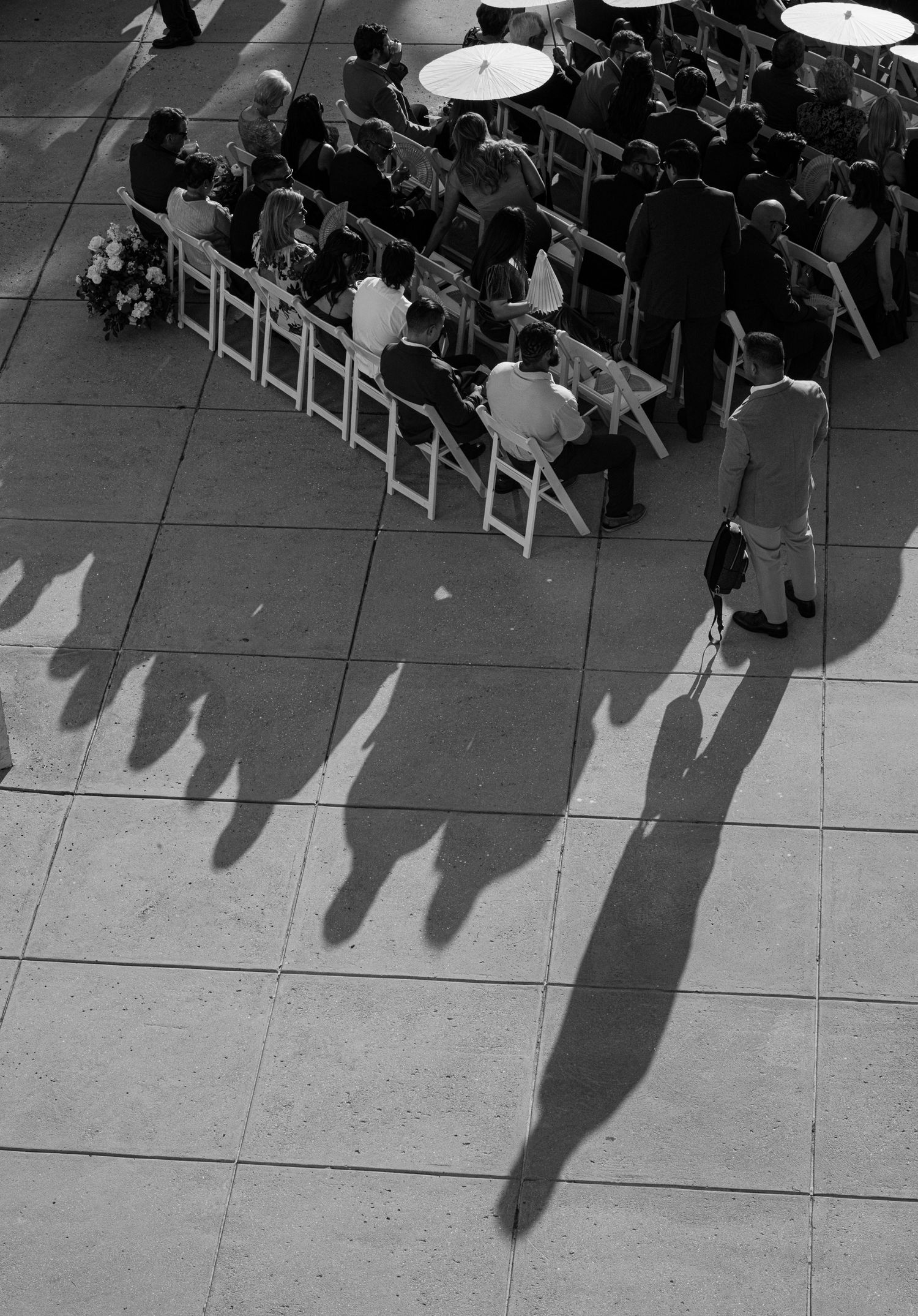 A black and white aerial view shows a line of seated people casting long dramatic shadows on a tiled floor.