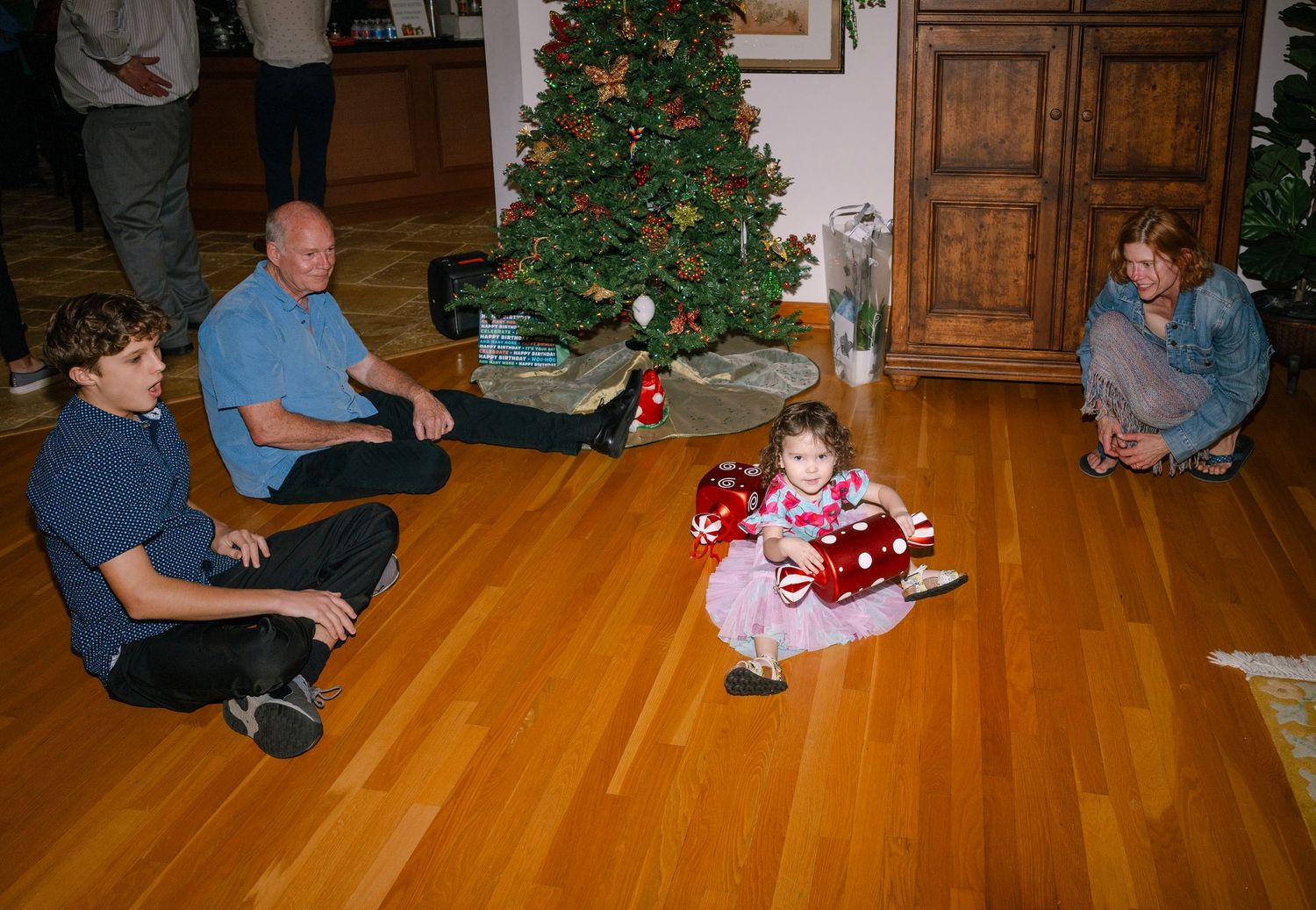 Family gathered around a decorated Christmas tree in a living room with hardwood floors during holiday gift opening.