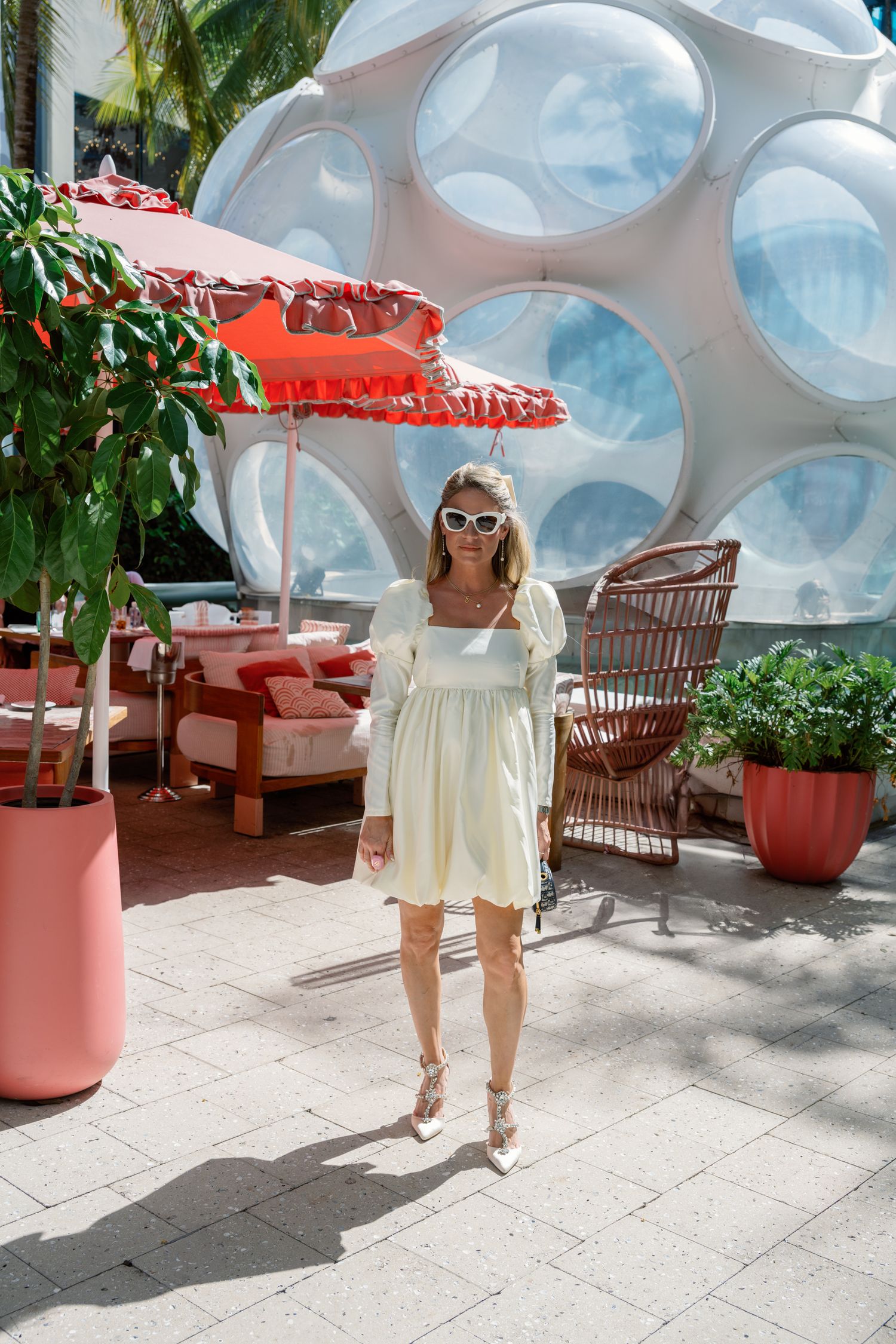 A person in a white summer dress and sunglasses walks along a modern patio with bubble-like architectural features.
