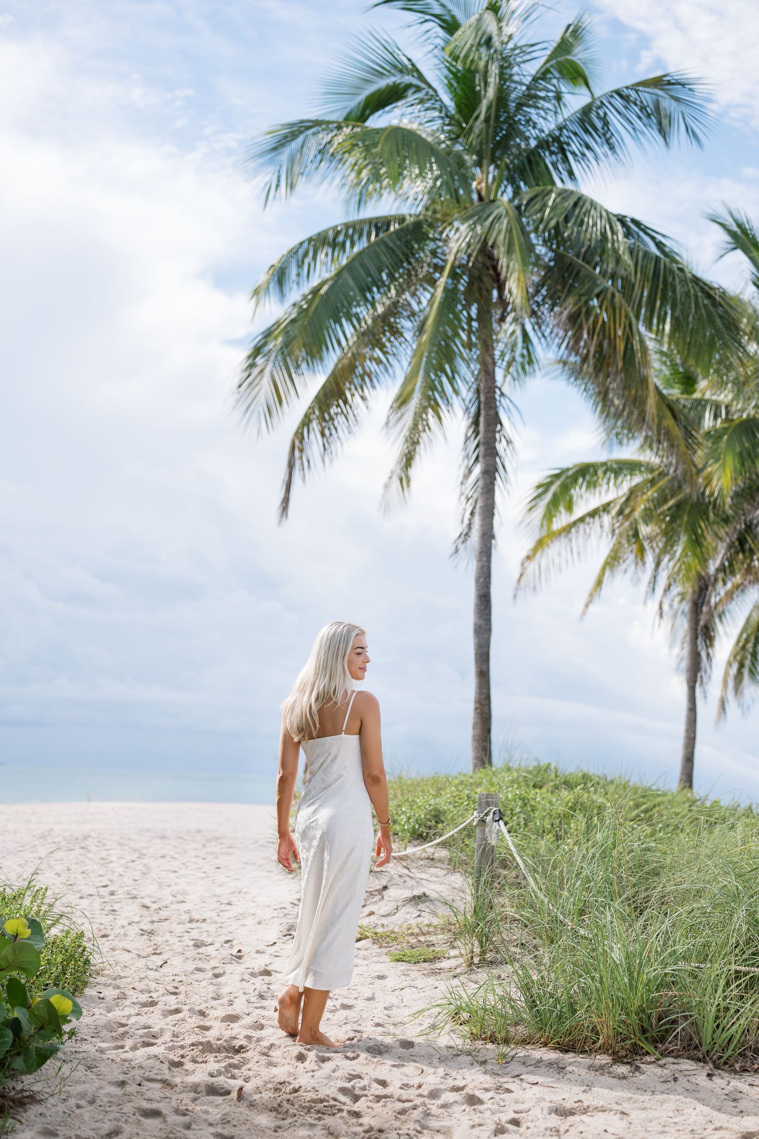 Woman in white dress walks along tropical beach path with palm trees and white sand on sunny day.