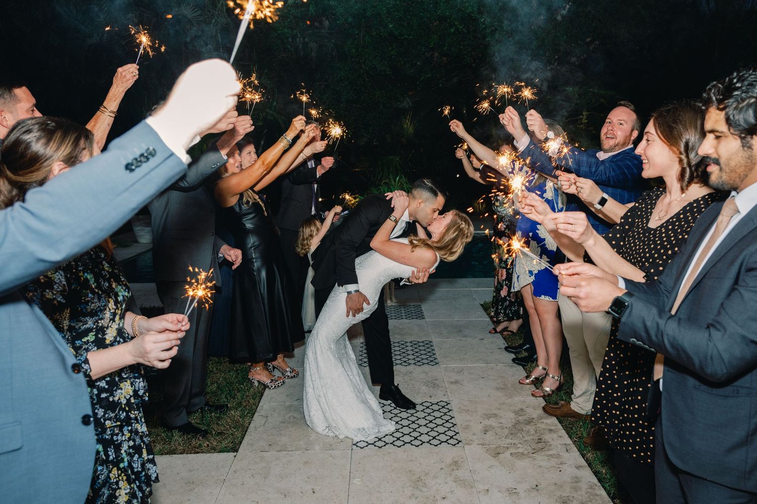 Wedding guests hold sparklers in the dark during a nighttime celebration at an outdoor reception venue.