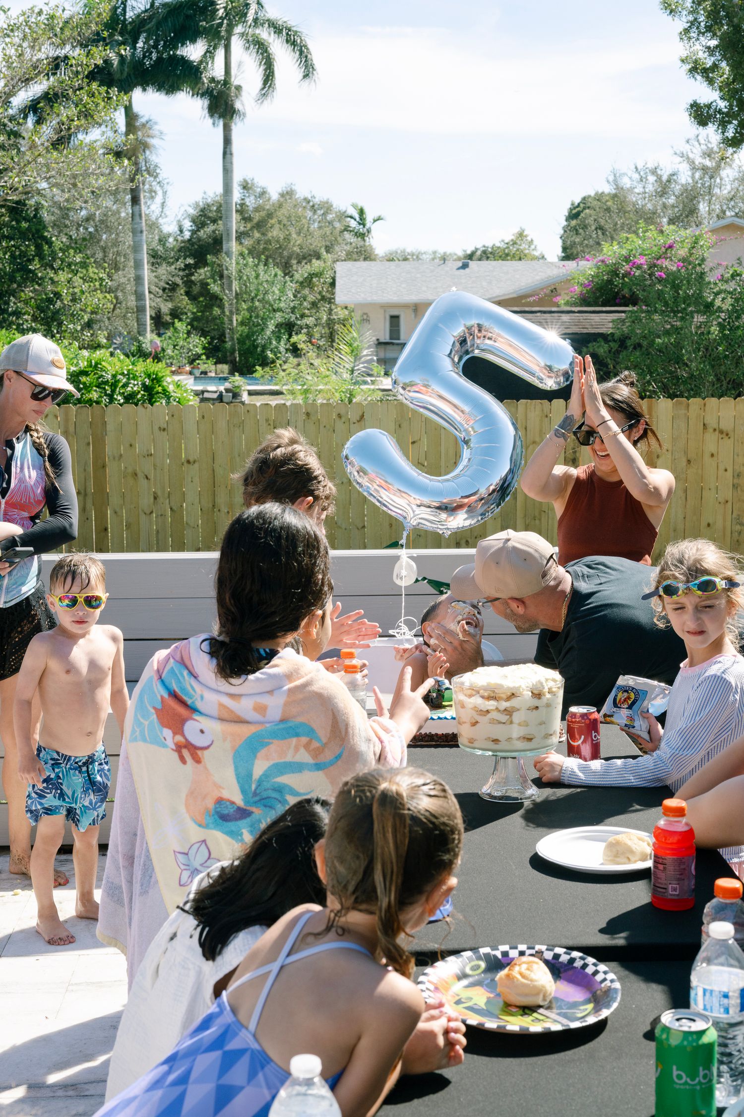 Outdoor birthday party celebration with blue number 5 balloon, cake and guests gathered around a table in a backyard.