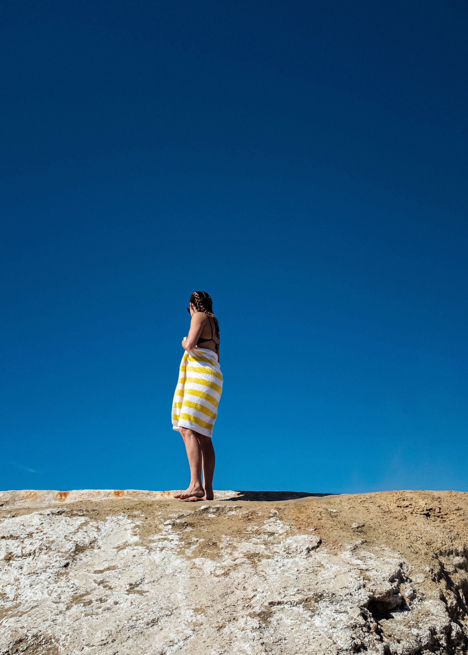 Woman wrapped in yellow striped towel standing on rocky cliff against clear blue sky.