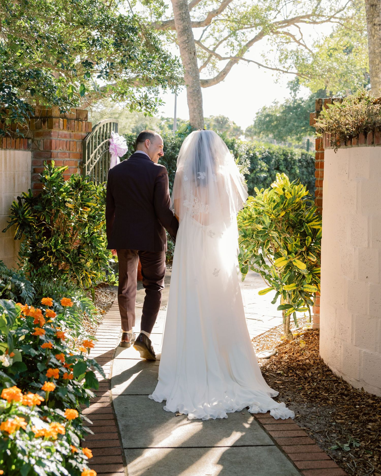 Couple walking along a sunlit garden path with white columns and orange flowering plants.