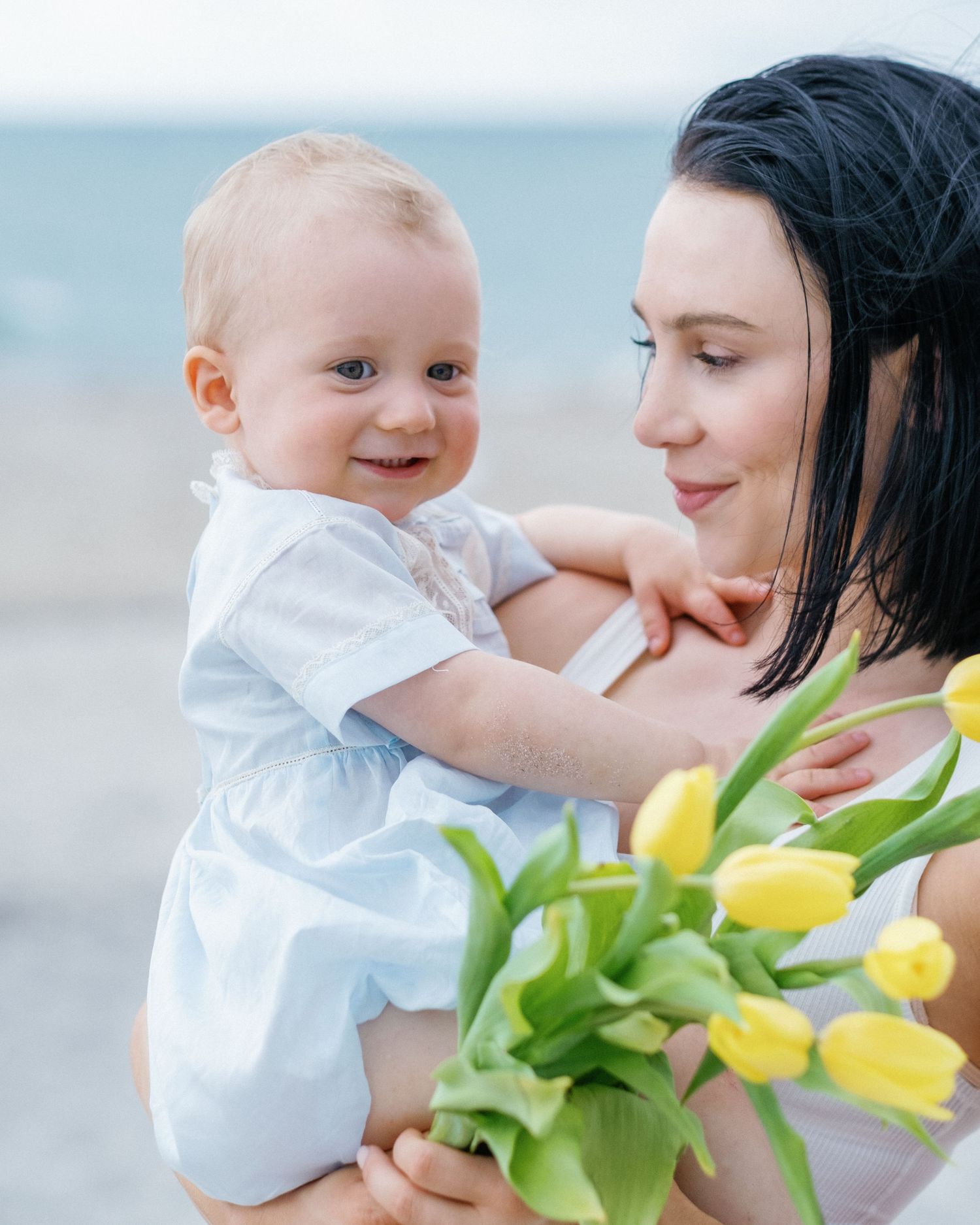 A tender moment between parent and baby in white clothing with yellow tulips on a beach.