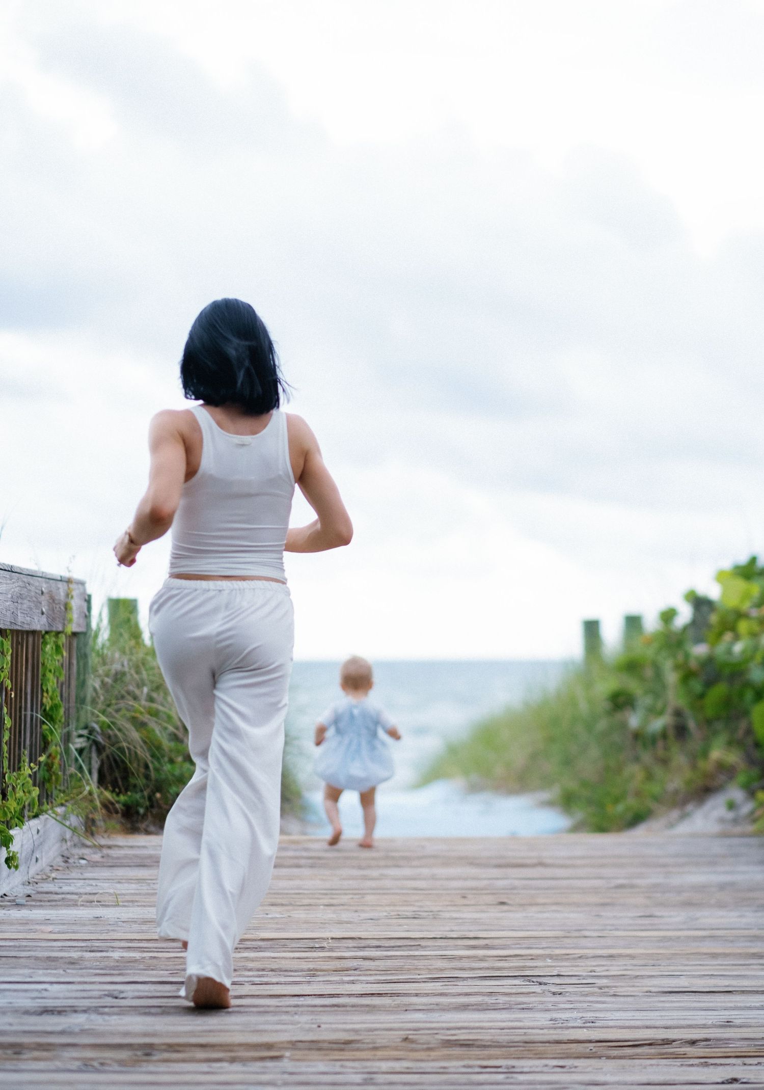 A person in white clothing walks along a wooden boardwalk path leading to the beach on a cloudy day.