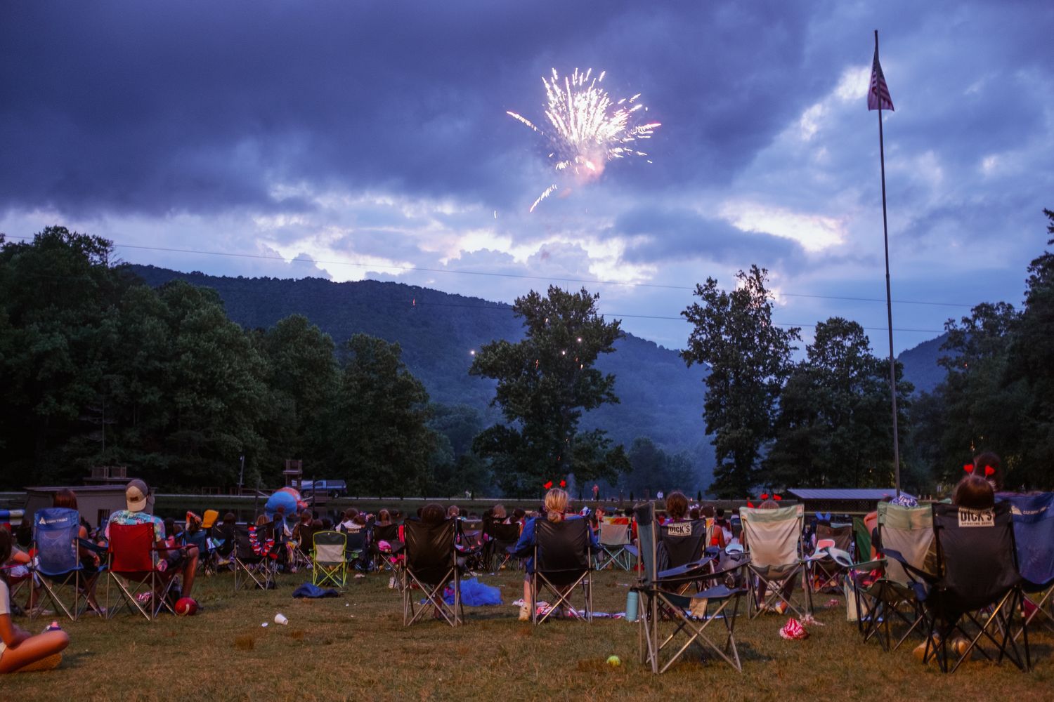 Fireworks illuminate a dark sky over a hillside campground with tents and RVs visible in the distance.