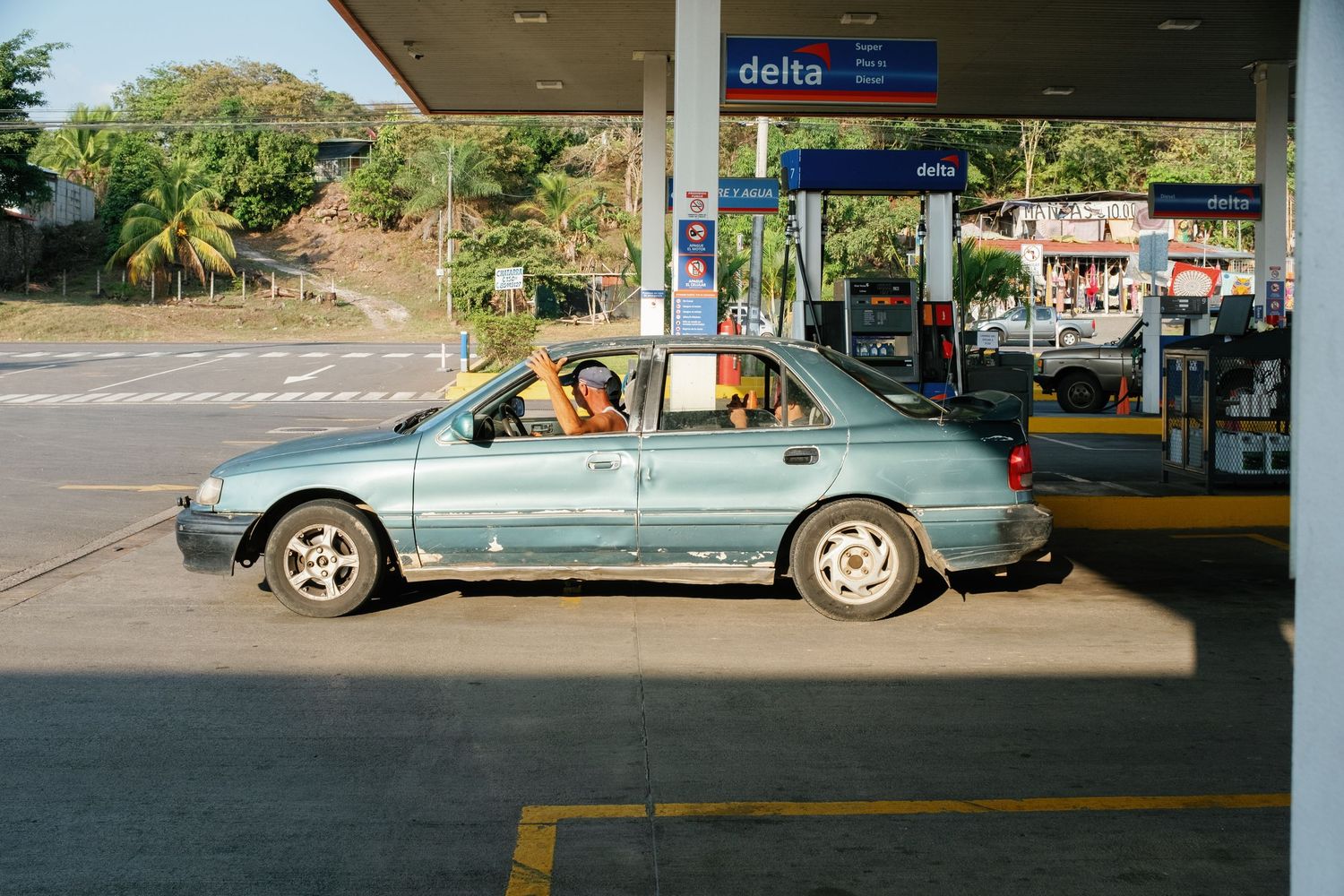 Light blue sedan refueling at a Delta gas station, driver visible through open window, tropical setting.