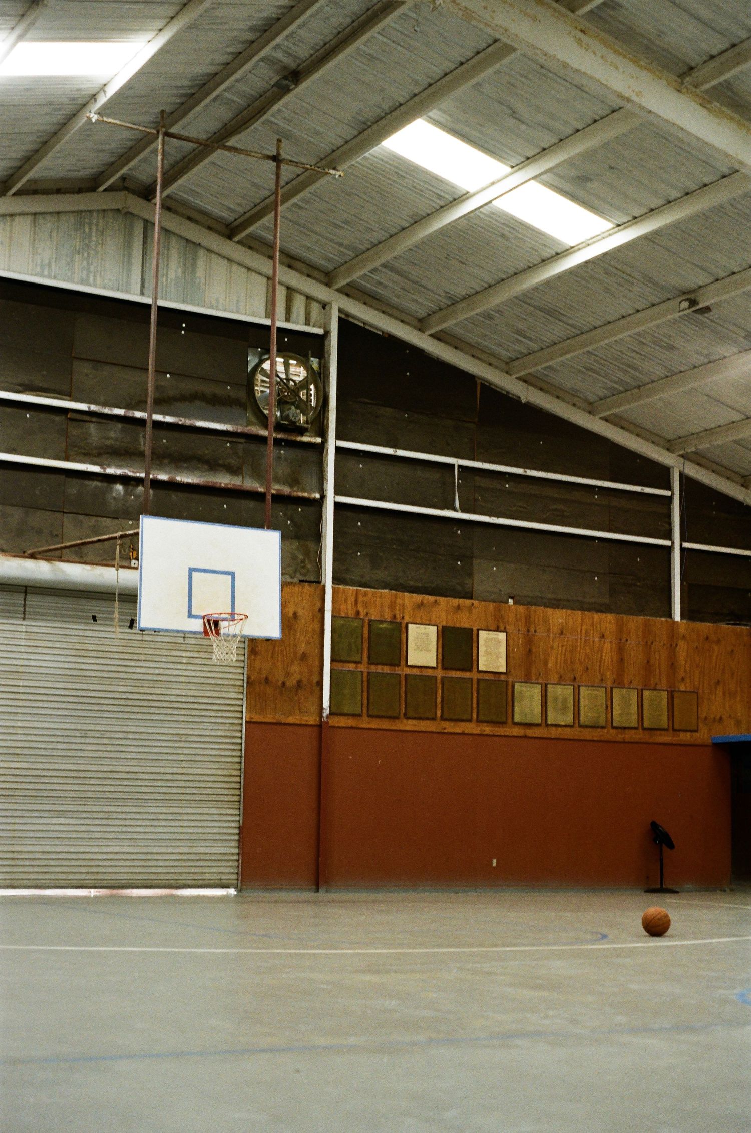 Indoor basketball court in a metal warehouse gym with a backboard, hoop, fan, wooden wall panels, and a basketball on the floor.