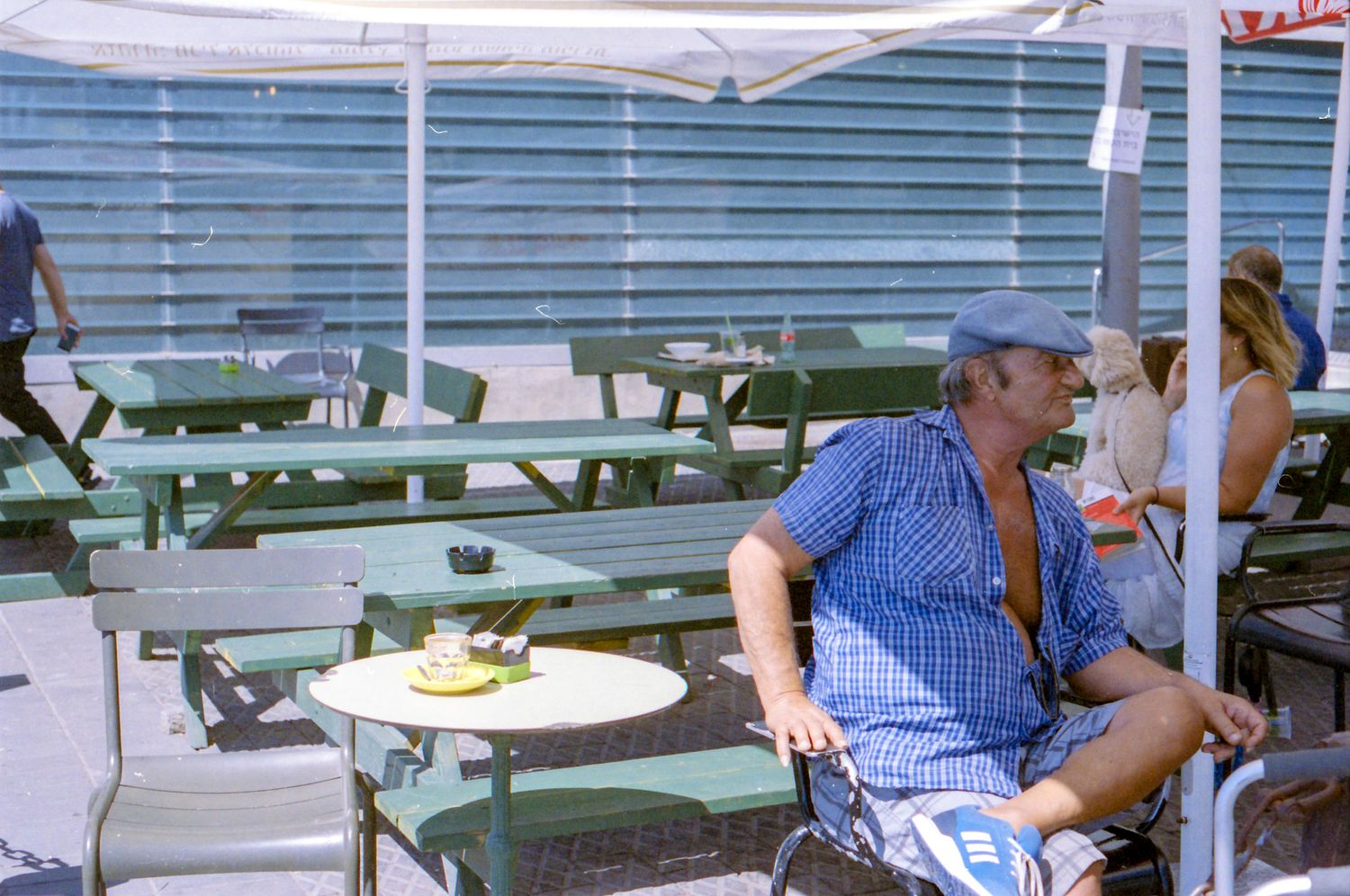People relaxing at outdoor seating area with green picnic tables and benches on sunny day.