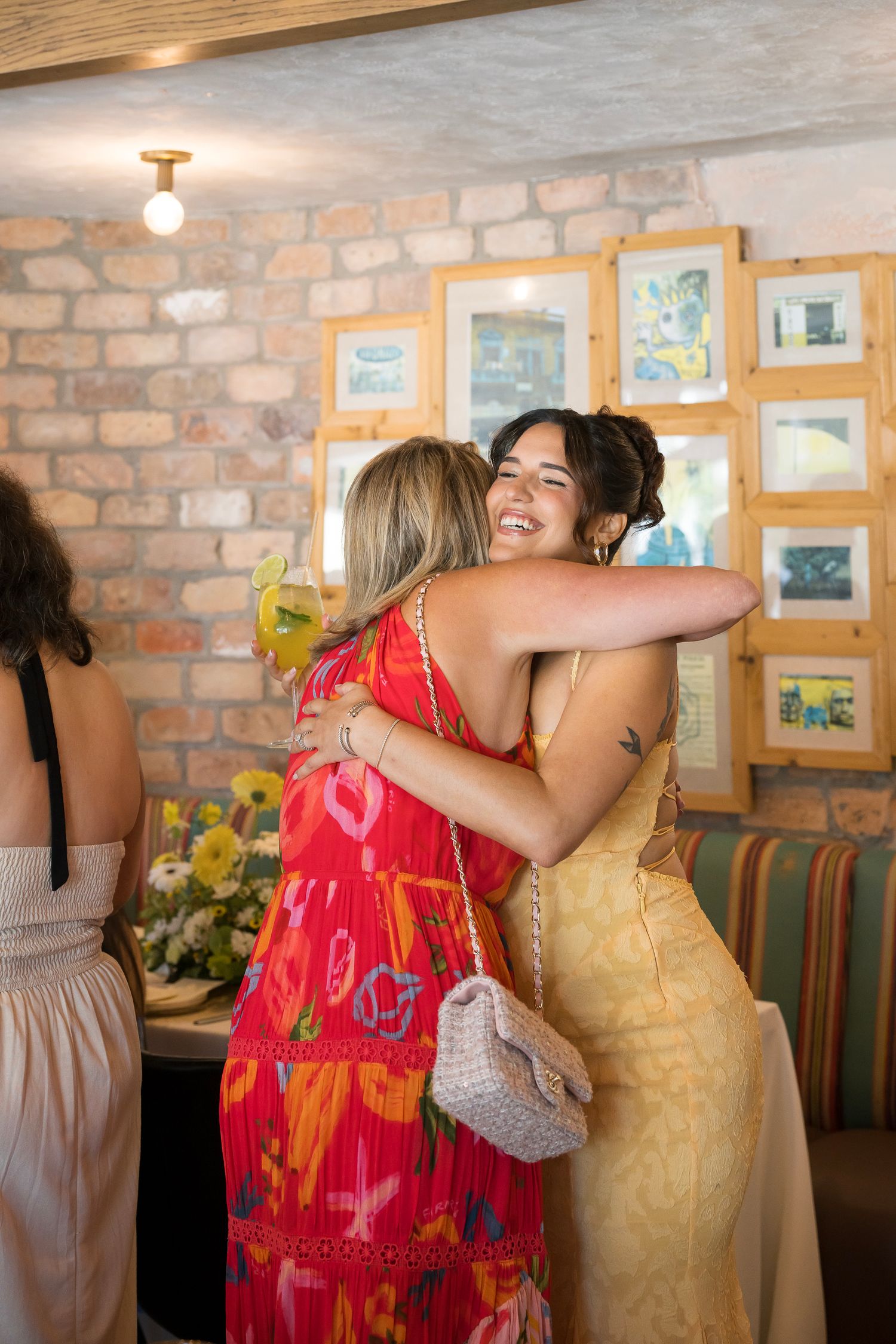 People in colorful dresses share warm embraces at a gathering with brick walls and artwork in the background.
