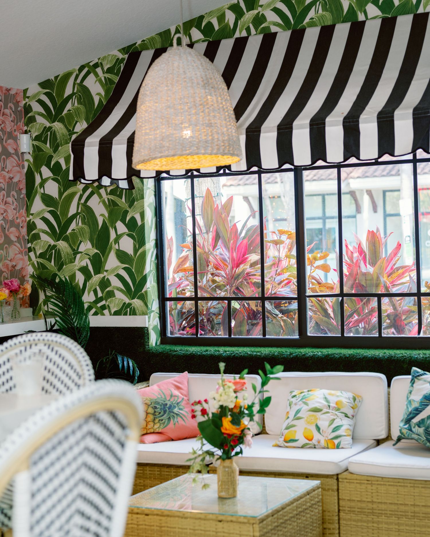 Bright restaurant interior with tropical leaf wallpaper, striped awning, and pendant lighting over banquette seating.