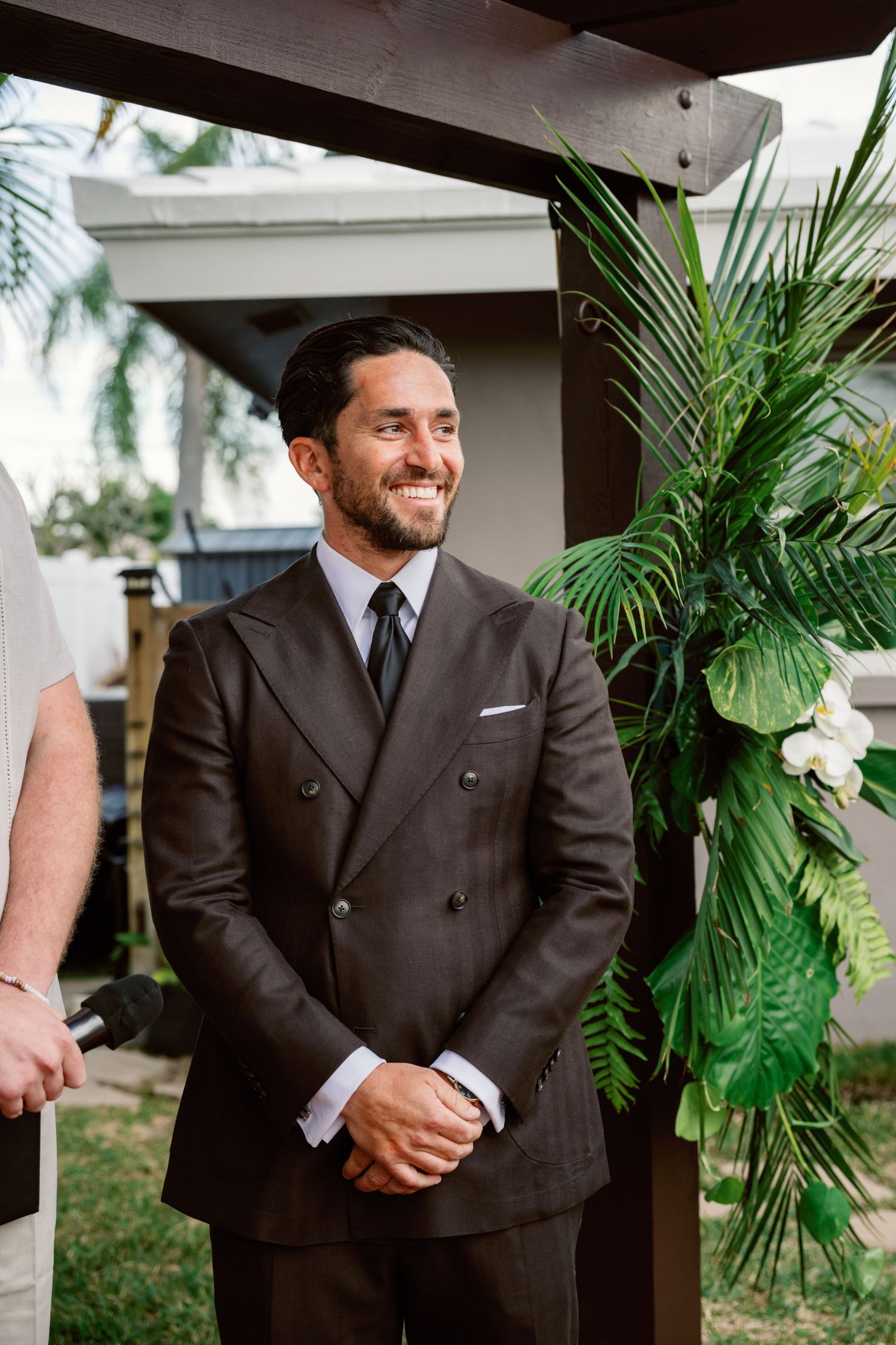 Groom in a dark suit stands against a backdrop of tropical palm plants at an outdoor wedding venue.