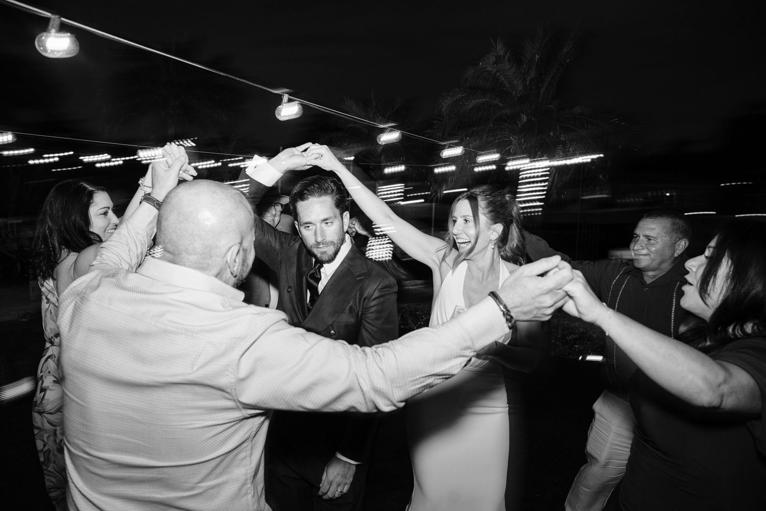 Wedding guests dancing and celebrating under string lights at a nighttime reception.