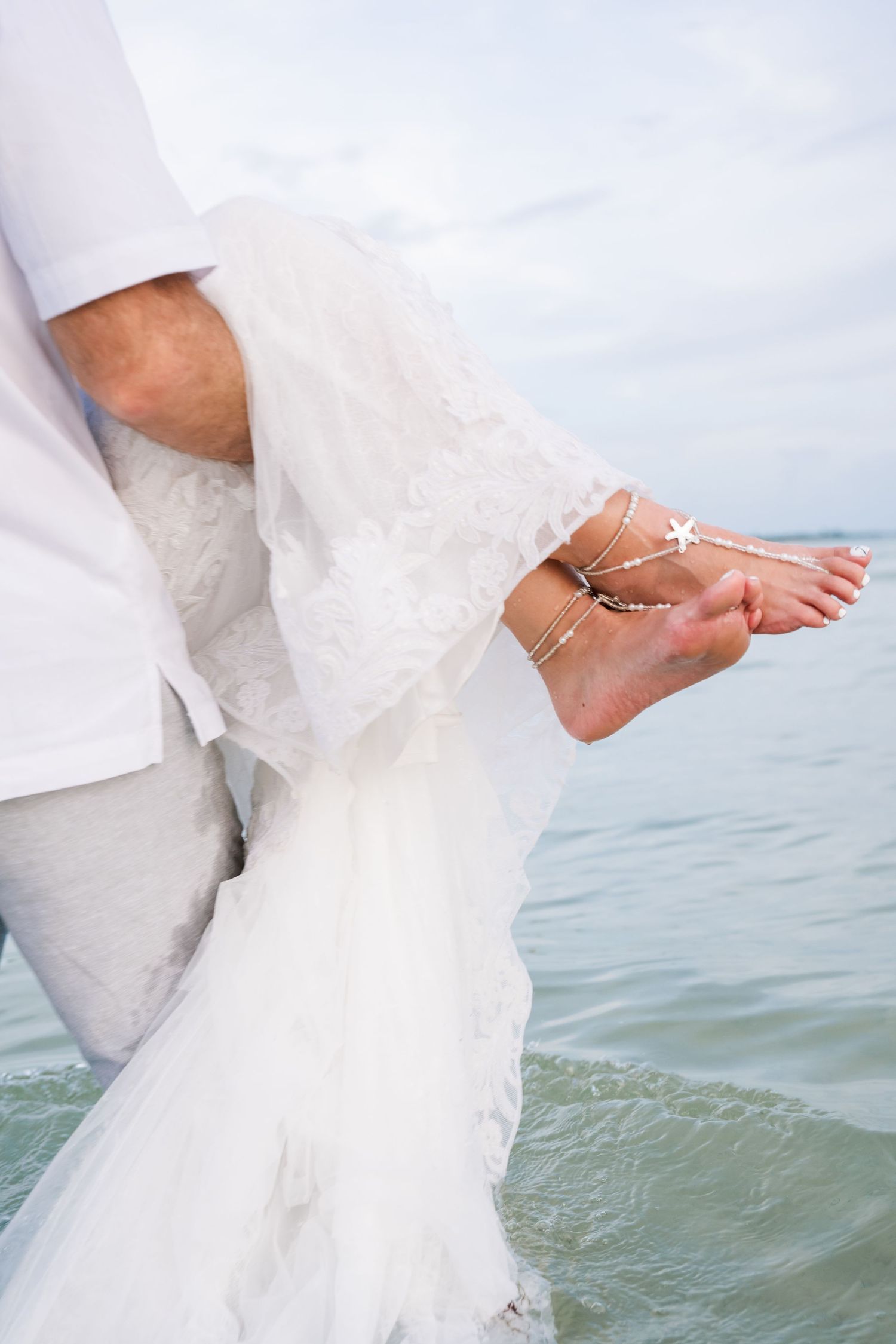 A bride's feet adorned with delicate anklets dip into gentle ocean waves while wearing a flowing white wedding dress.