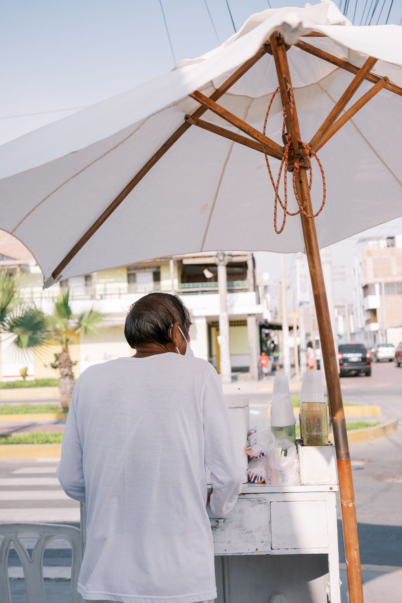 Street vendor in white shirt working at outdoor cart under large white umbrella on a sunny urban street.