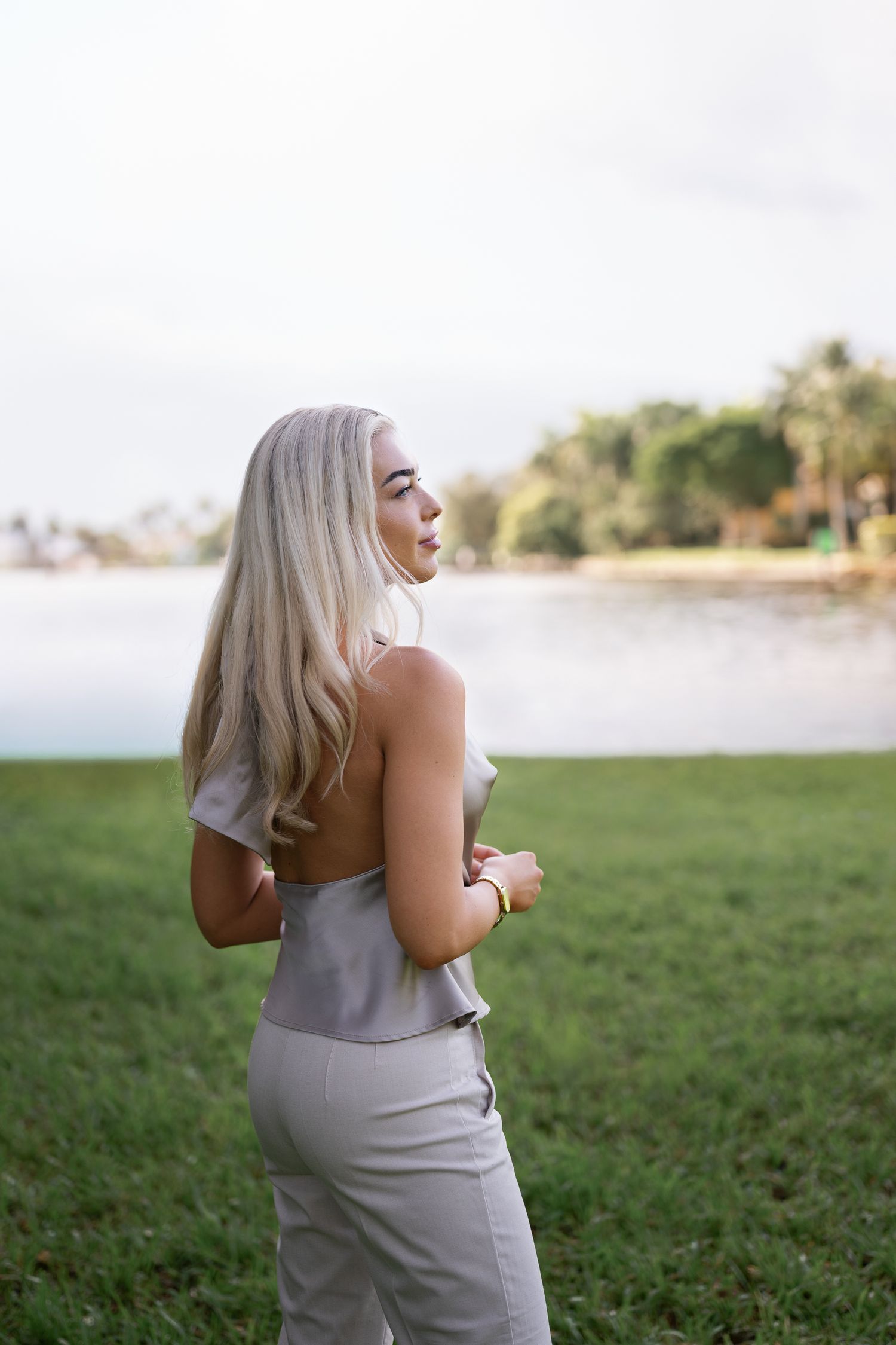 Woman with long platinum blonde hair in a sleek grey dress stands on grass by a tranquil lake at sunset.
