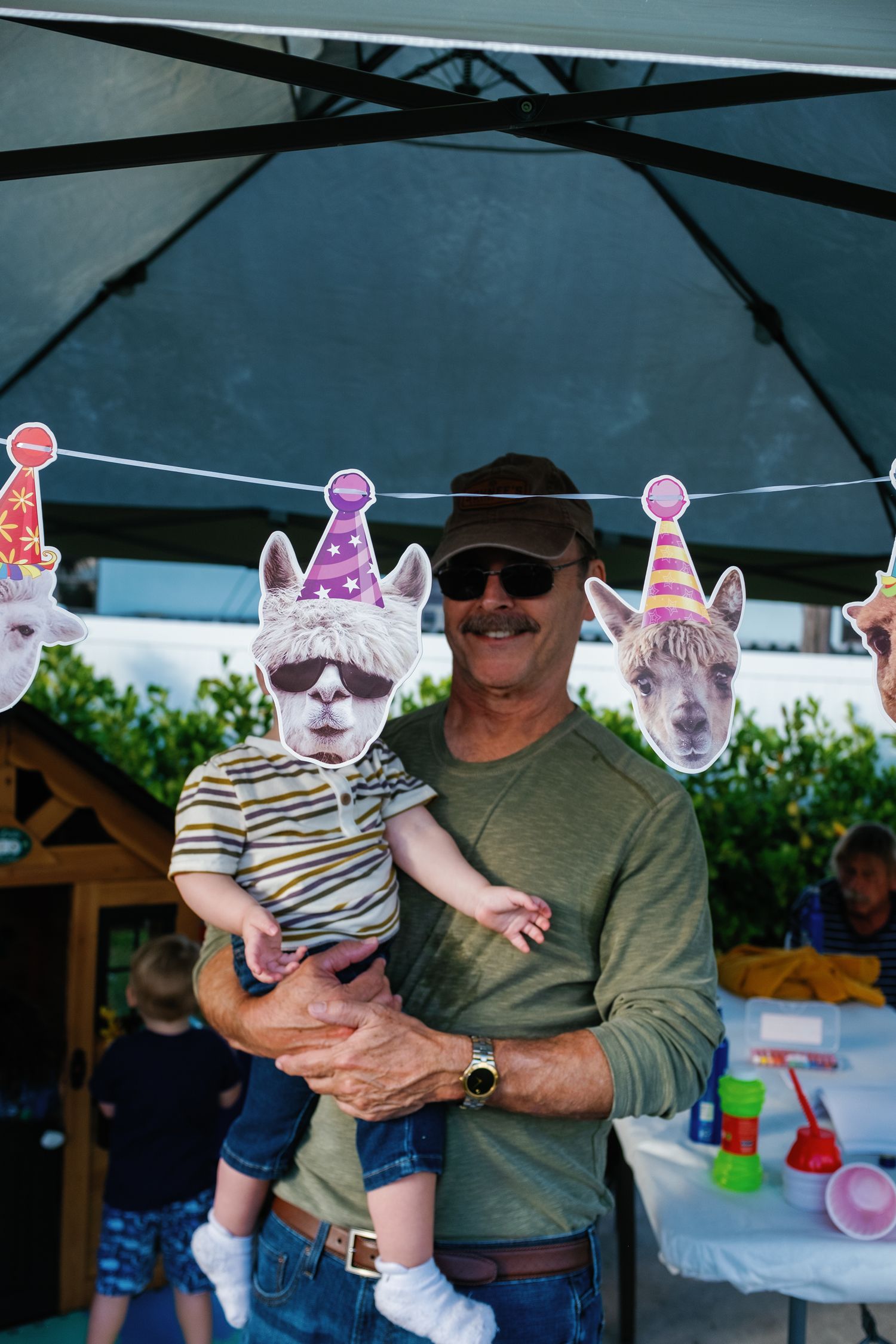 A series of photos showing a baby wearing party accessories being held under a blue umbrella at an outdoor celebration.