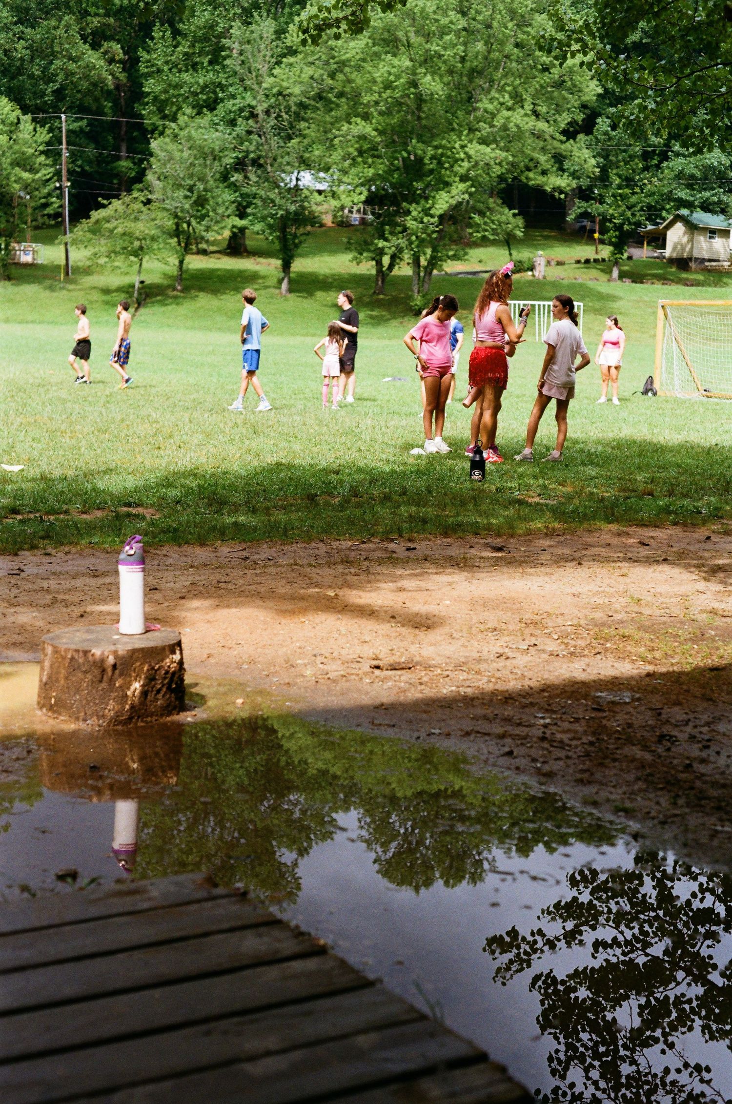 Children playing outdoor games on a grassy field near a pond at a summer camp, surrounded by lush green trees.