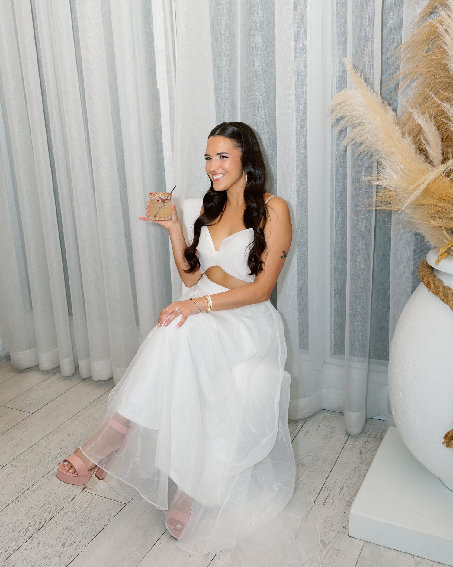 A person in a white tulle dress sits on a white floor with pampas grass decor in the background.