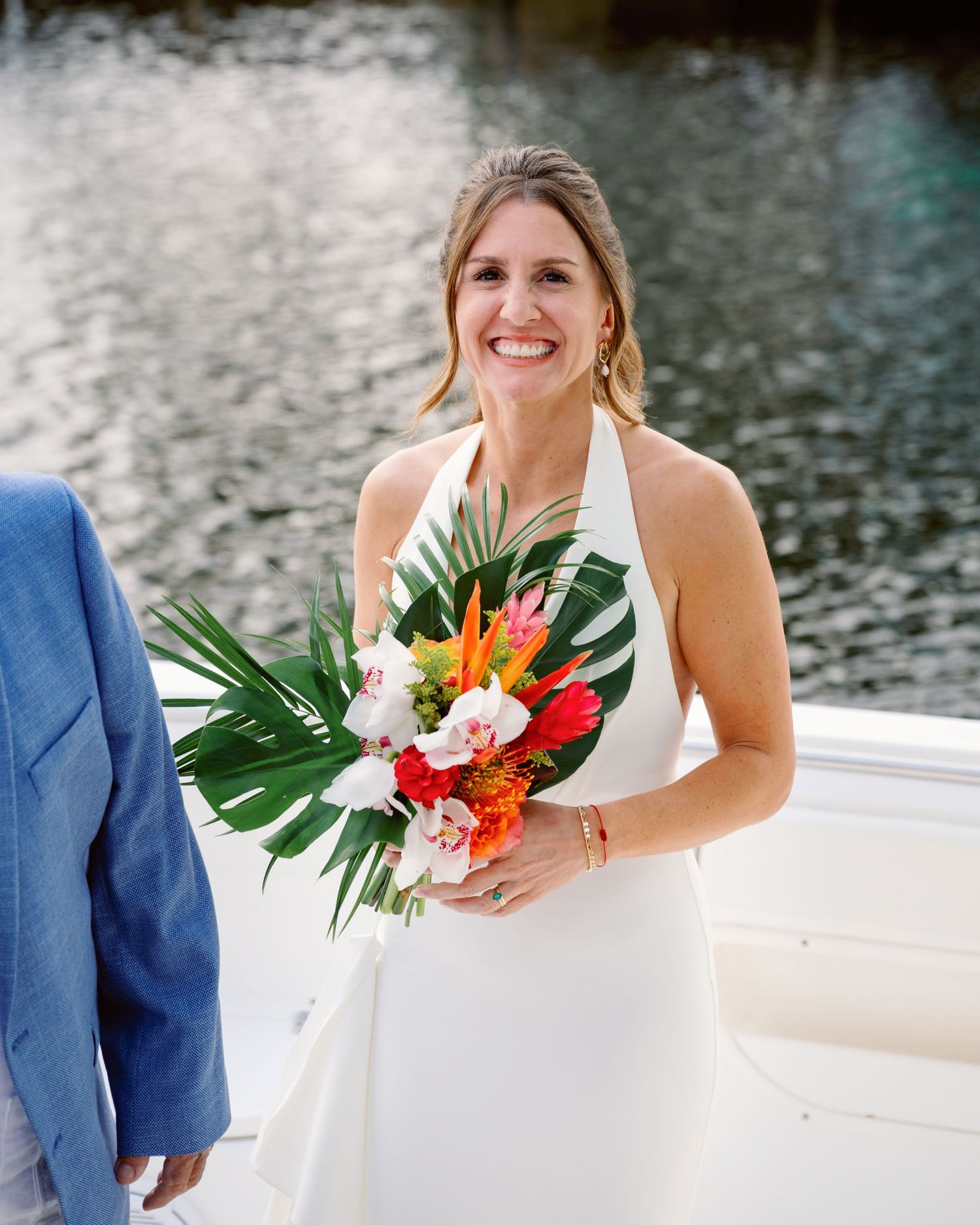 A tropical wedding bouquet with birds of paradise and palm leaves rests against a white dress by the water.